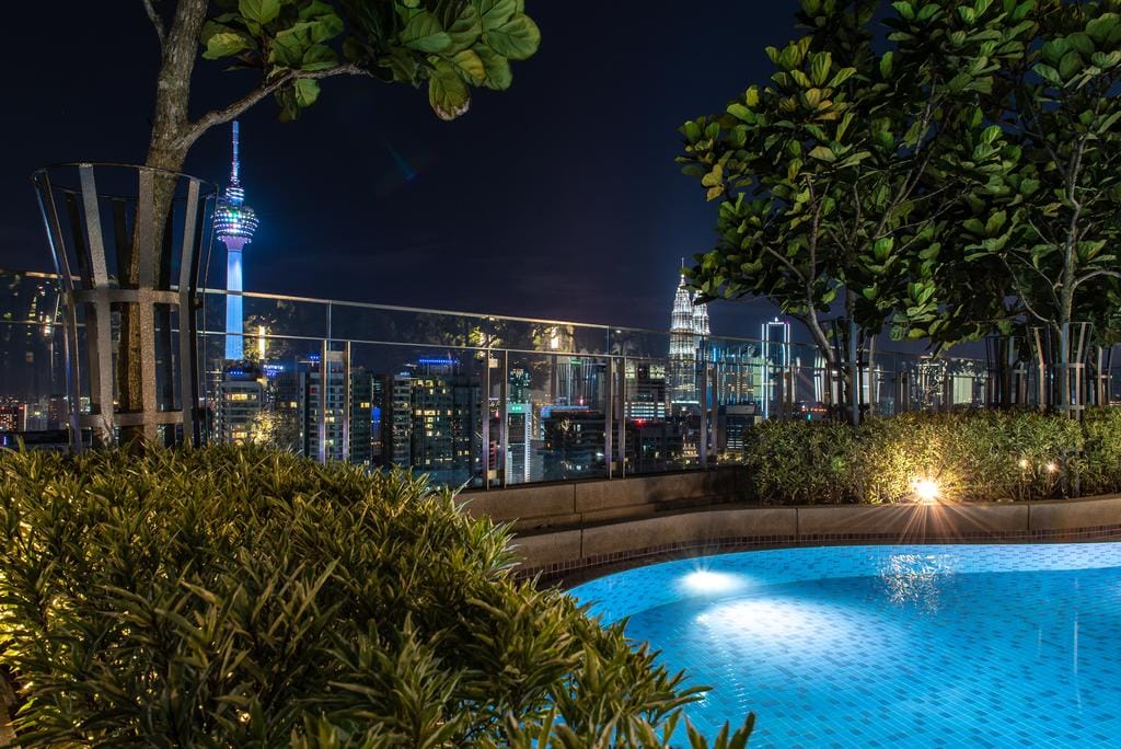 The rooftop area features a tranquil infinity pool surrounded by lush greenery and subtle lighting. In the background, the KL Tower and city skyline are visible, creating a serene atmosphere against the night sky.
