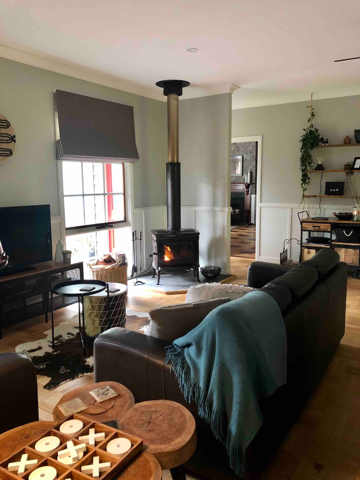 A cozy living area features a wood burner fireplace centered in the room, providing warmth and comfort. A leather sofa is complemented by a soft throw blanket, while a coffee table adorned with decorative coasters and wooden trays adds charm. A television is positioned nearby, with natural light streaming through a window.