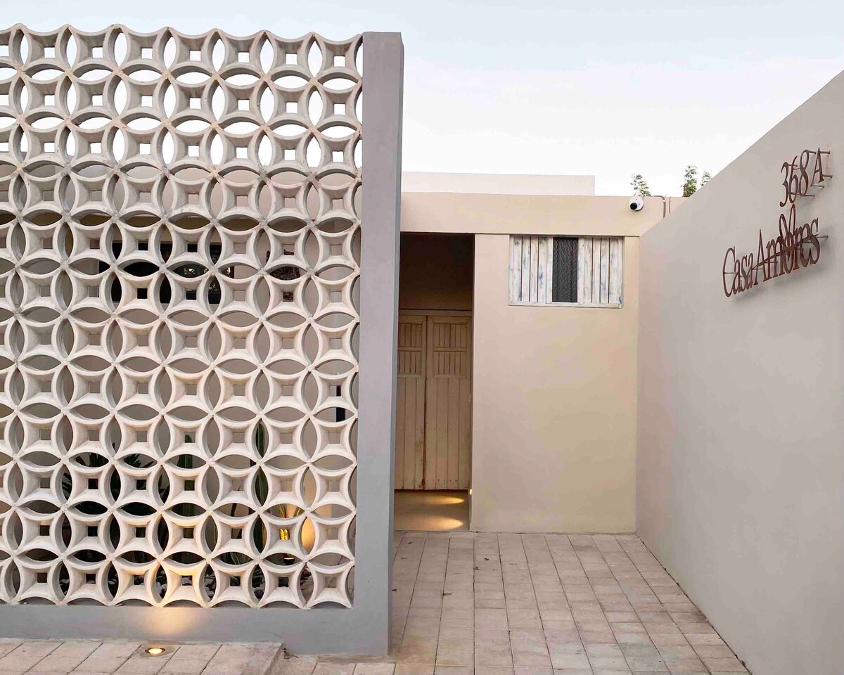 The exterior of Casa Amores is highlighted by a unique decorative wall feature, showcasing a geometric design. The entrance is framed by neutral-colored plaster, and a subtle sign indicates the property name. Soft lighting accentuates the path leading to the entry door.
