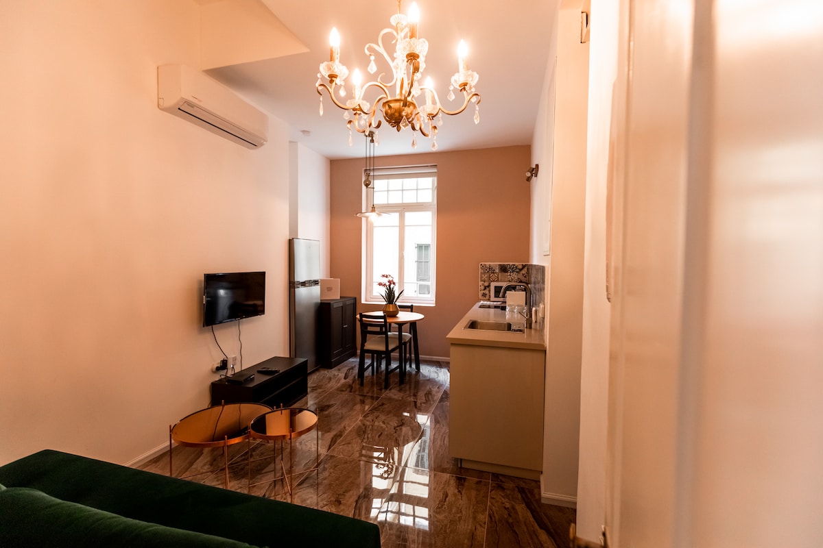 A stylish living area is highlighted by a green sofa bed and marble flooring. A decorative chandelier hangs from the ceiling, while a dining table for two sits near a window, allowing natural light to fill the room. Modern appliances are arranged in the kitchenette.