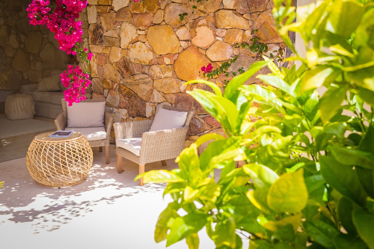 A serene courtyard setting features a natural stone wall adorned with vibrant pink flowers. Two light-toned armchairs are positioned beside a woven coffee table, creating a relaxing seating area surrounded by lush green foliage.