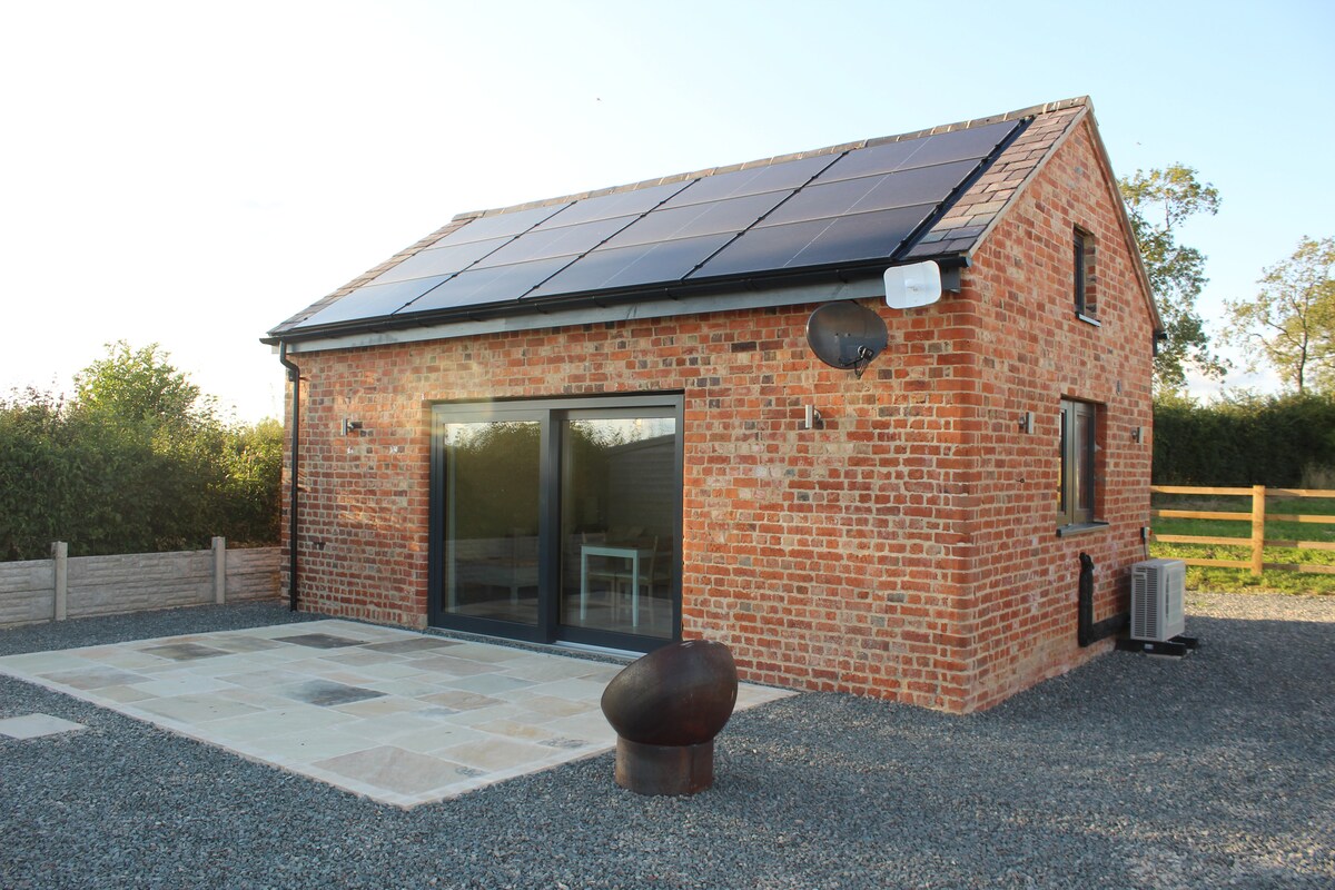 The exterior of a brick barn conversion is showcased, featuring solar panels on the roof and large glass doors that provide natural light. A stone patio area is visible, accompanied by a modern sculpture and gravel landscaping, set against a backdrop of greenery.