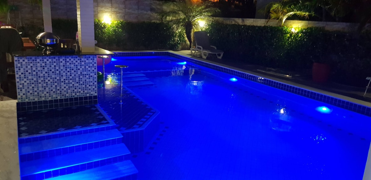 A large pool is illuminated in various shades of blue, creating a calming atmosphere. Steps leading into the water are visible, as well as lounge chairs positioned nearby. Soft lighting highlights the surrounding greenery and the tiled surfaces.