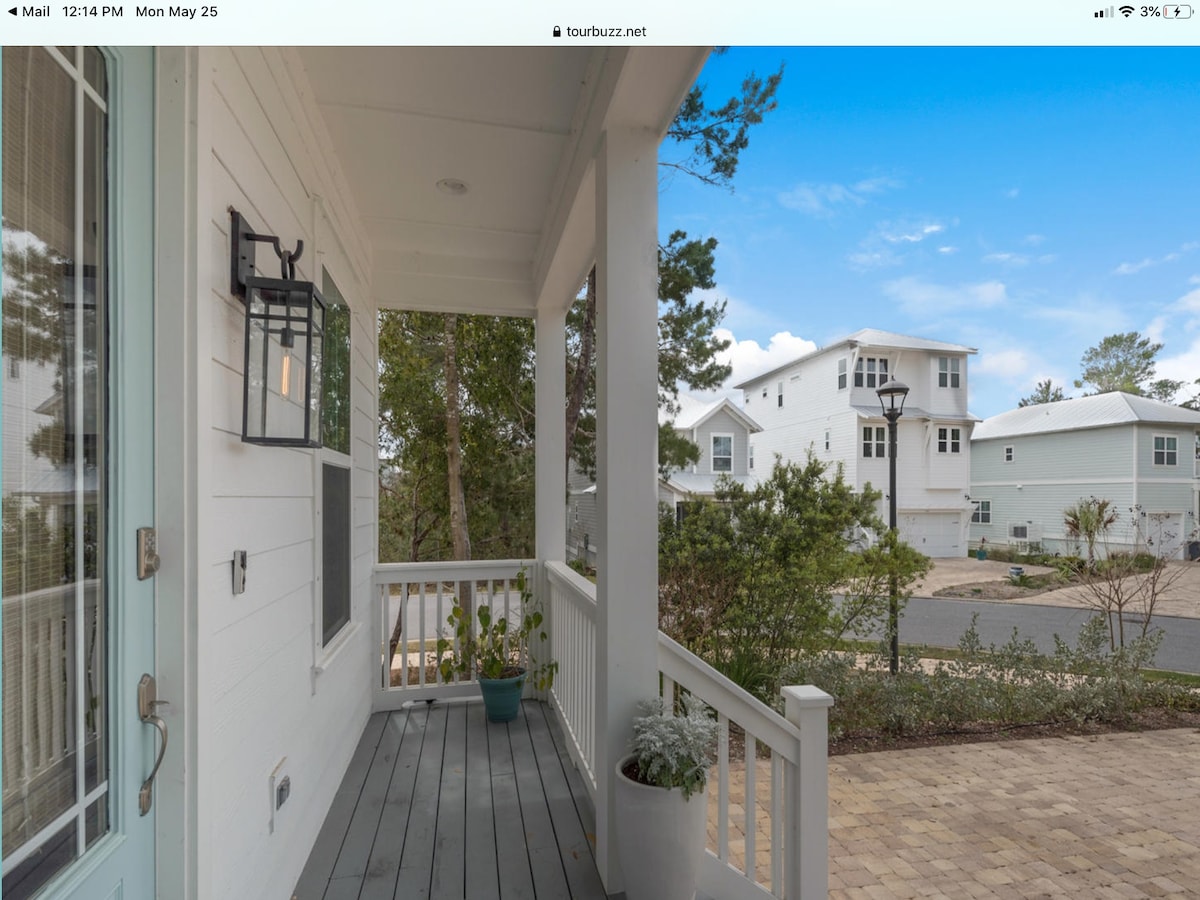 The front porch is depicted with a wooden floor and light-colored railing. A potted plant is positioned near the entrance, while natural greenery borders the walkway. Nearby homes are visible in the background, along with a clear sky, which enhances the inviting exterior setting.