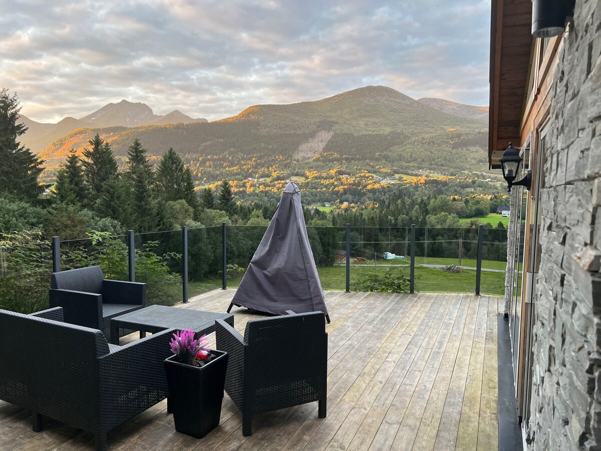A spacious deck features dark wicker seating arranged around a small table. A potted plant adds color to the area. The surrounding mountains and trees can be seen under a softly illuminated sky, creating a serene outdoor setting.
