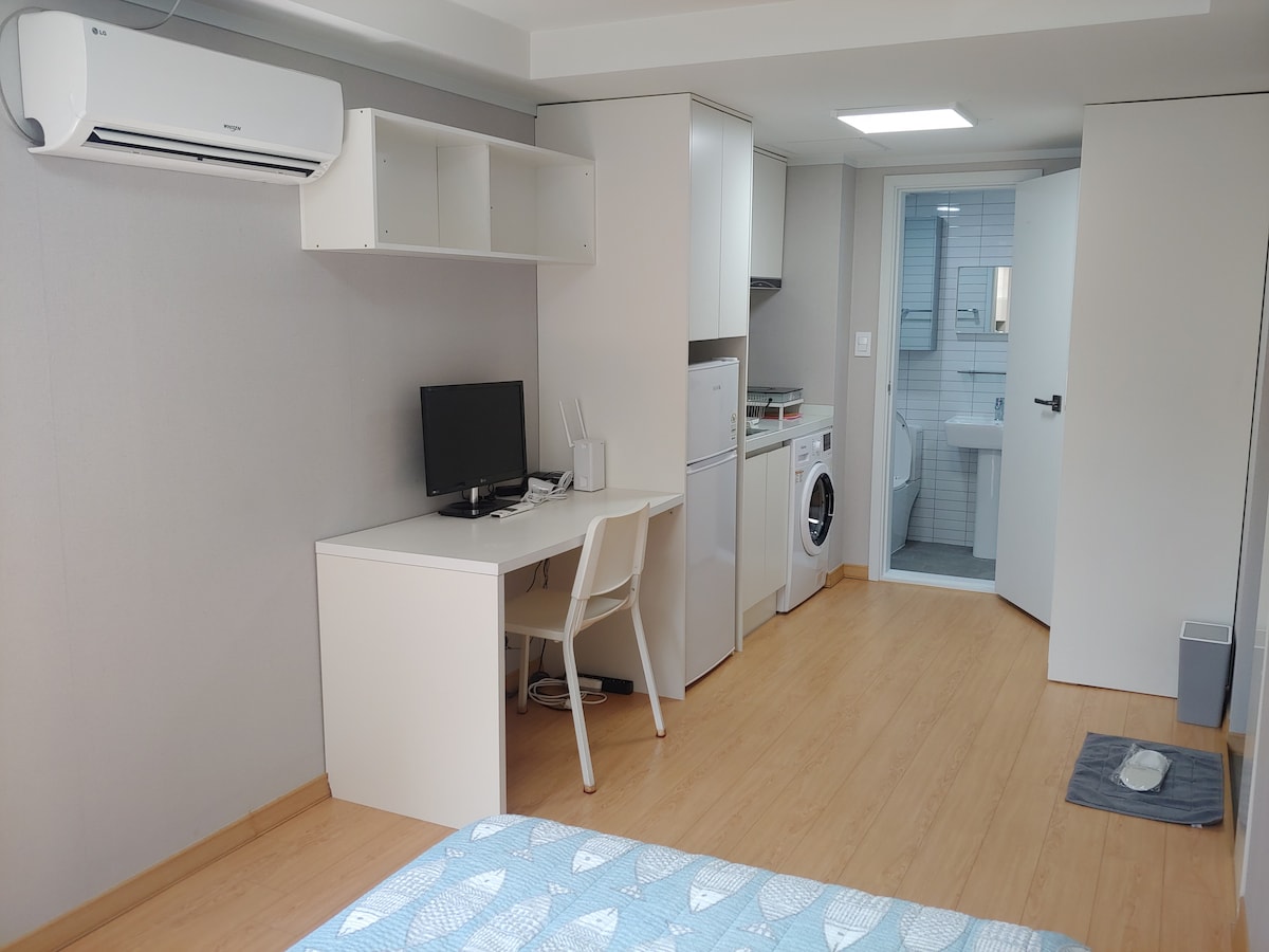 A studio room is depicted featuring modern furnishings. A desk with a computer is positioned near a washing machine, while a compact kitchen area contains a refrigerator and microwave. Bright wooden flooring enhances the spaciousness, and a doorway leads to a bathroom.