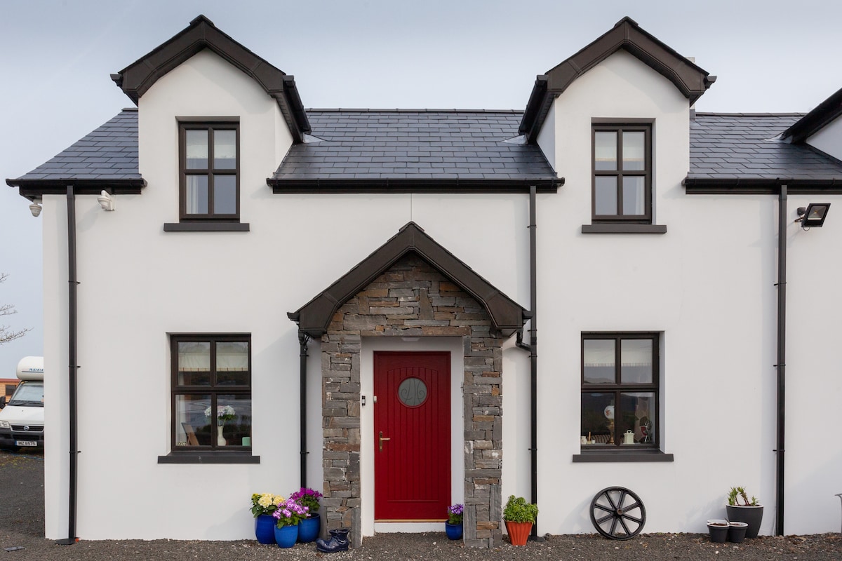 The exterior of the cottage features a white façade with a stone accent around the entrance. A bright red door is centrally located, flanked by windows on either side. Potted flowers in blue and terracotta containers add a touch of color to the entryway.