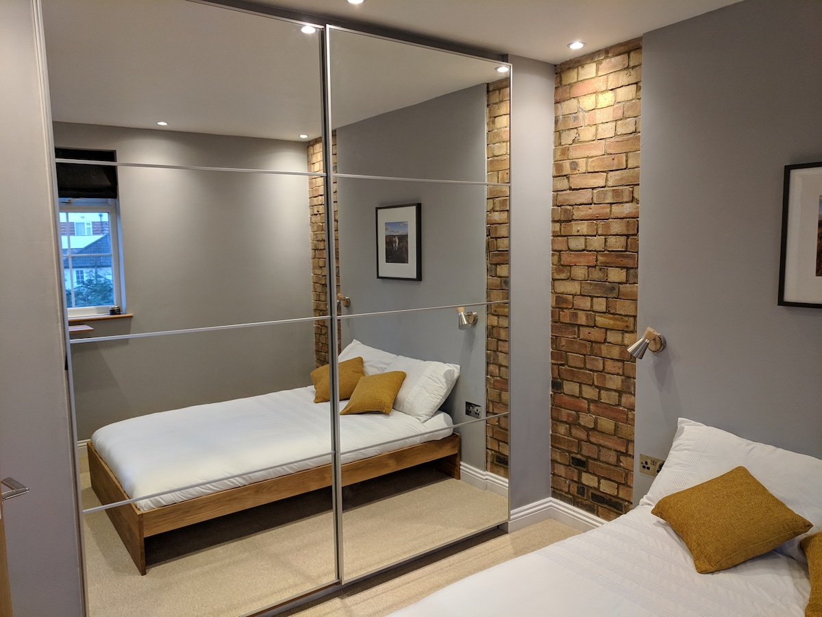 A modern bedroom features a spacious bed with a white duvet and a decorative pillow. Exposed brick walls add character, while a large mirrored wardrobe reflects the light. A window allows natural light to brighten the room, complemented by soft overhead lighting.