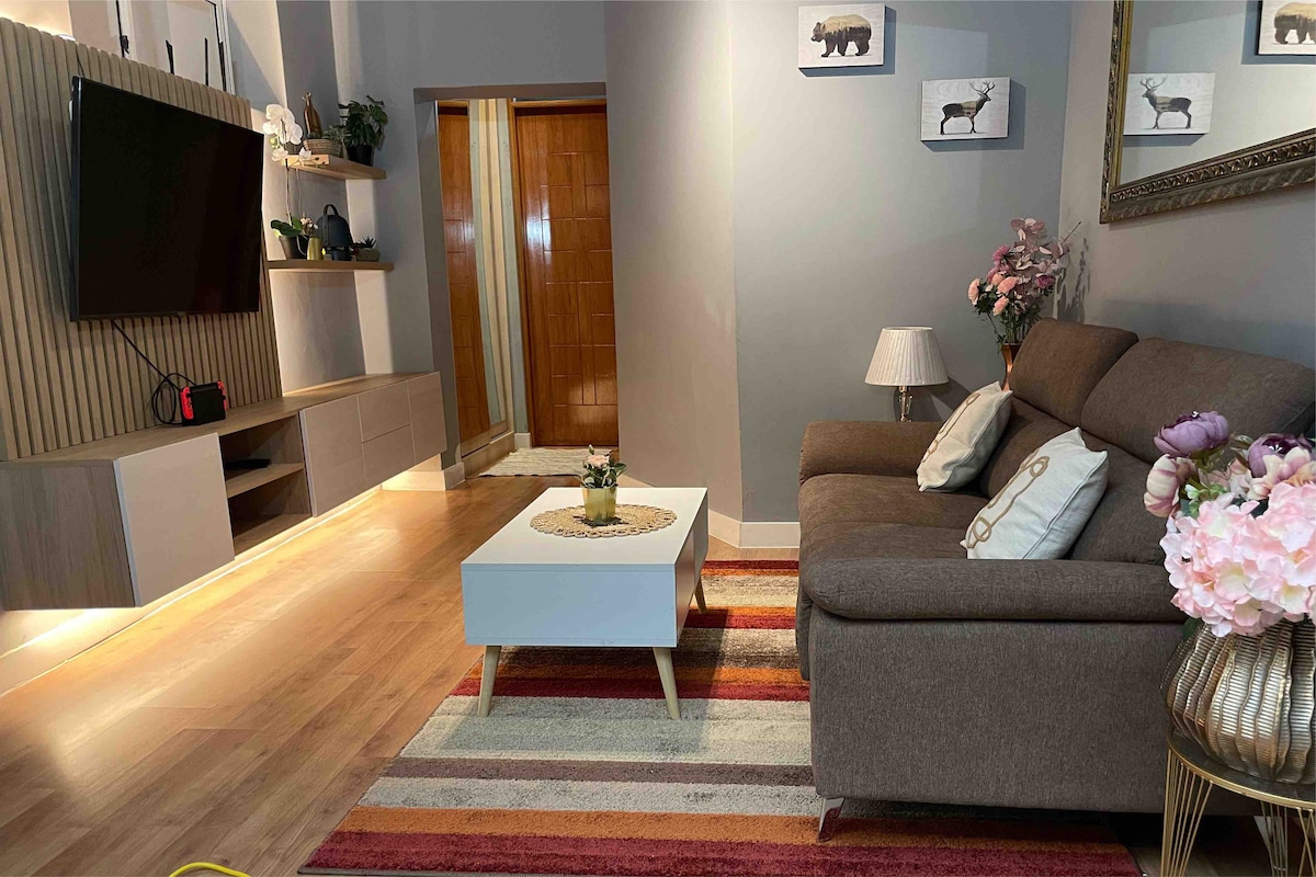 A living area is featured with a comfortable brown sofa and a white coffee table. A television is mounted on the wall, while decorative plants and a lamp provide soft lighting. Colorful rugs enhance the space, complementing the light wood flooring.