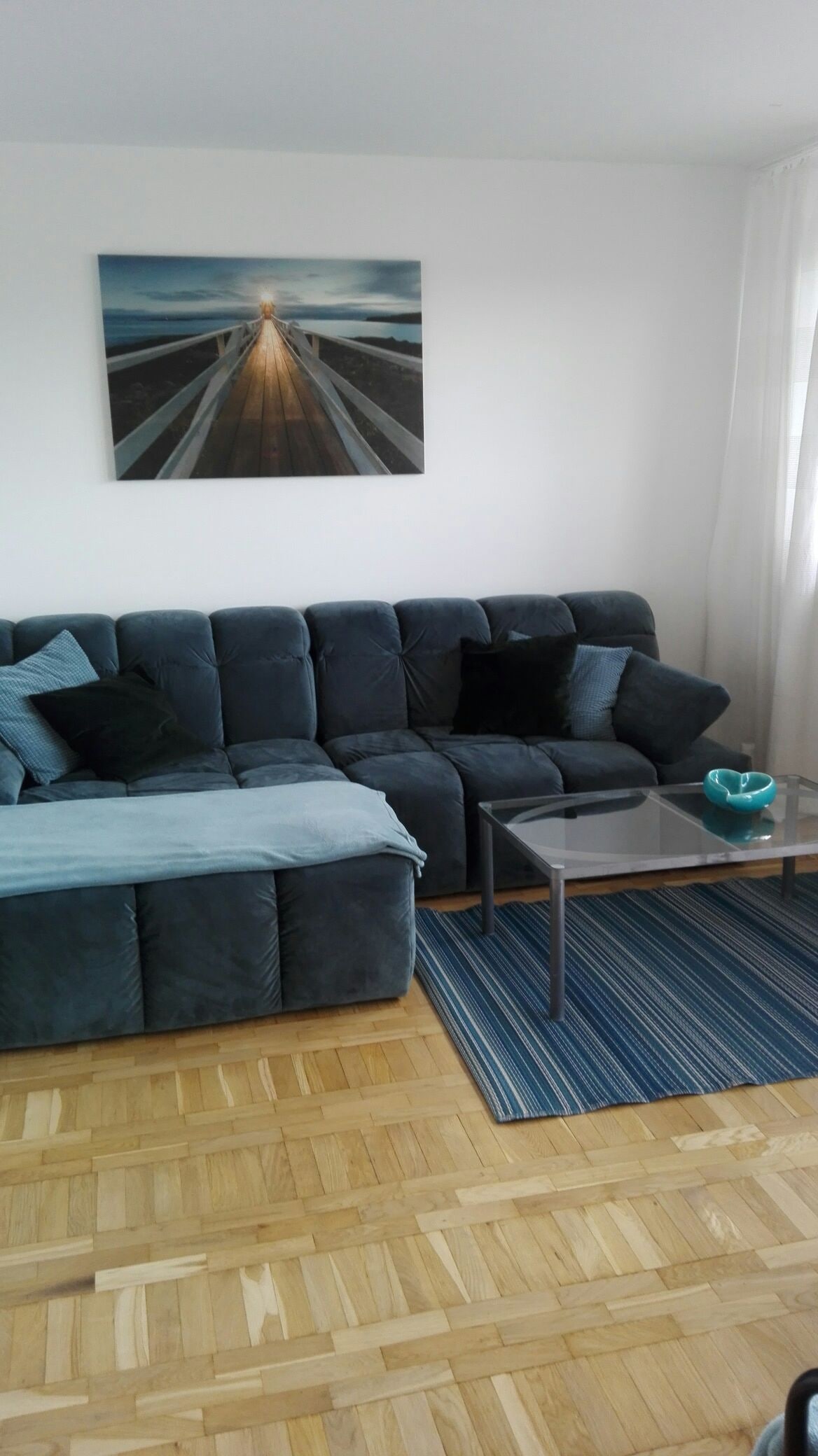 A spacious living area features a soft grey sectional sofa adorned with blue and black pillows. A compact glass coffee table rests on a blue striped rug, while natural light enters through large windows framed by sheer white curtains. A serene wall art piece complements the décor.