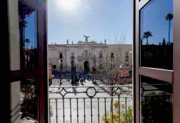 A view from the balcony features a bustling plaza with a historic building at the center. The scene is framed by open wooden windows, allowing natural light to illuminate the space. Palm trees are visible in the background, enhancing the outdoor ambiance.