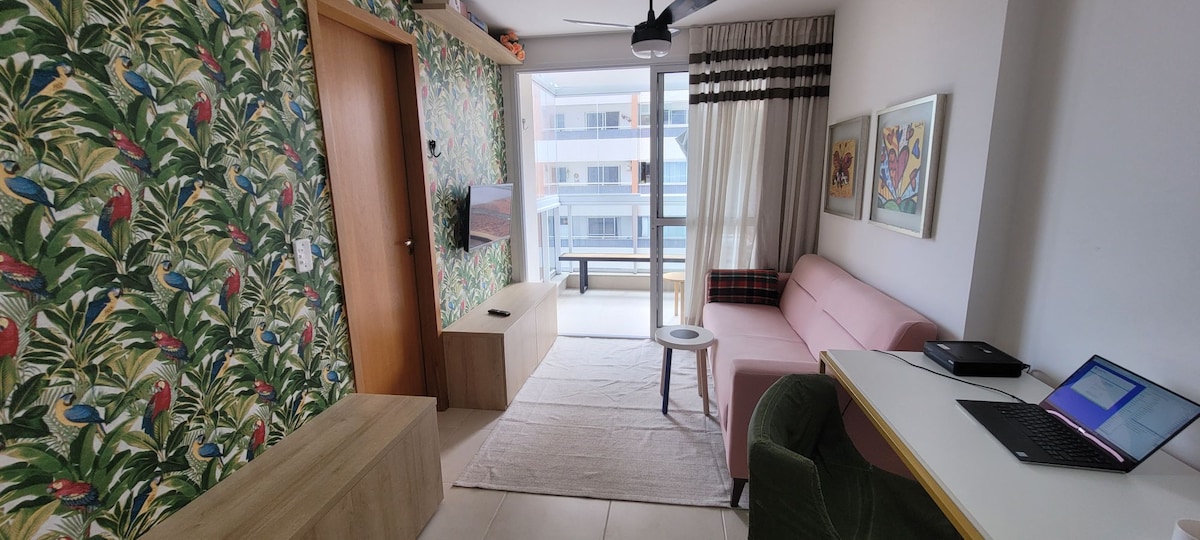 A vibrant living area is adorned with tropical-themed wallpaper featuring greenery and colorful birds. A pink sofa and a green armchair provide comfortable seating. A small table stands next to the sofa, while a work desk with a laptop is positioned against the wall.