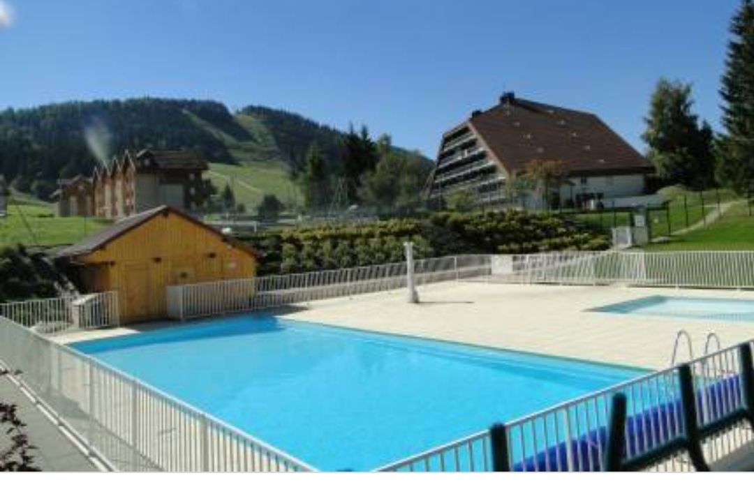 An outdoor swimming pool is presented, featuring a clear blue water surface and a surrounding deck area. Lush green hills rise in the background, along with a multi-story building and several smaller cabin-like structures. Fencing encloses the pool area for safety.