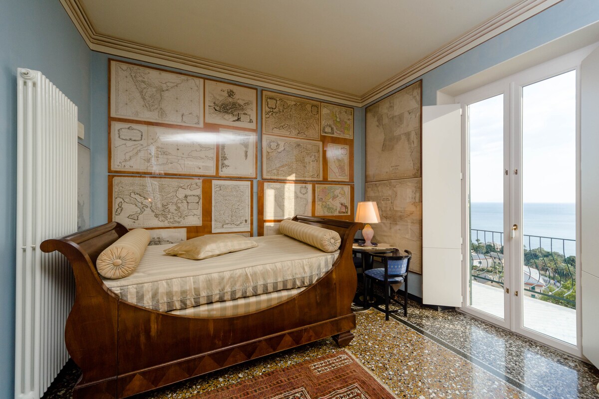 A spacious sleeping area is presented with a curved wooden bed positioned against walls adorned with vintage maps. Natural light enters through large doors that open to a sea view. A bedside table with a lamp complements the setting, adding functionality.