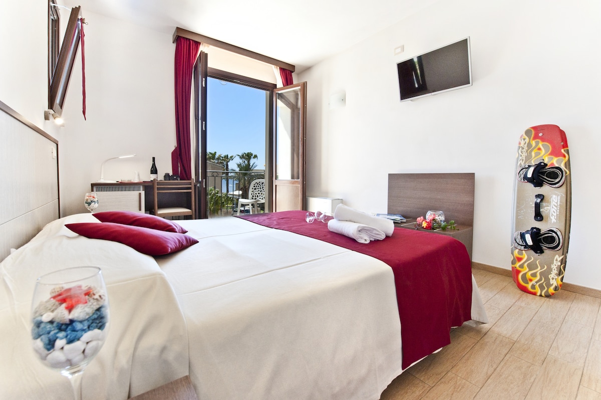 A light-filled room features a double bed dressed in white linens with a deep red throw, complemented by a second bed across the space. A small desk and chair are positioned beside a window that opens to outdoor views. A television is mounted on the wall.
