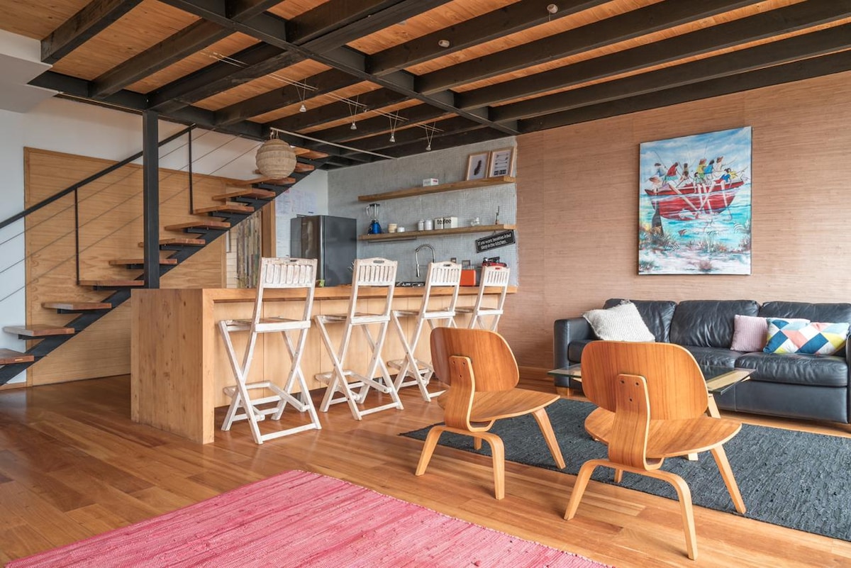 An open living space features a modern kitchen island with high stools and a cozy seating area. Light wood flooring contrasts with the dark wooden beams overhead. A colorful artwork adorns the wall, while a warm textile rug provides a comfortable visual base.