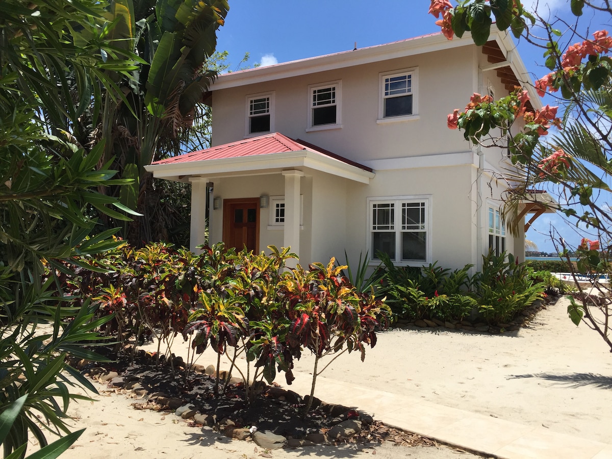 The exterior of Ginger House is presented, showcasing a modern two-story structure with a red roof. Lush greenery and colorful flowering plants surround the entrance, while a sandy pathway leads to the front door. The beach is visible in the background.