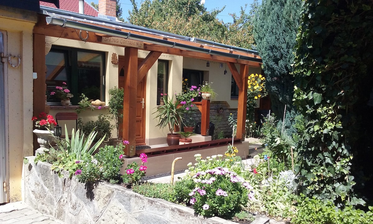 The exterior of the cottage is framed by vibrant flower beds and greenery. A wooden porch features potted plants and yellow flowers, providing a welcoming view. The entrance is surrounded by trees, enhancing the cottage's charming and private garden setting.