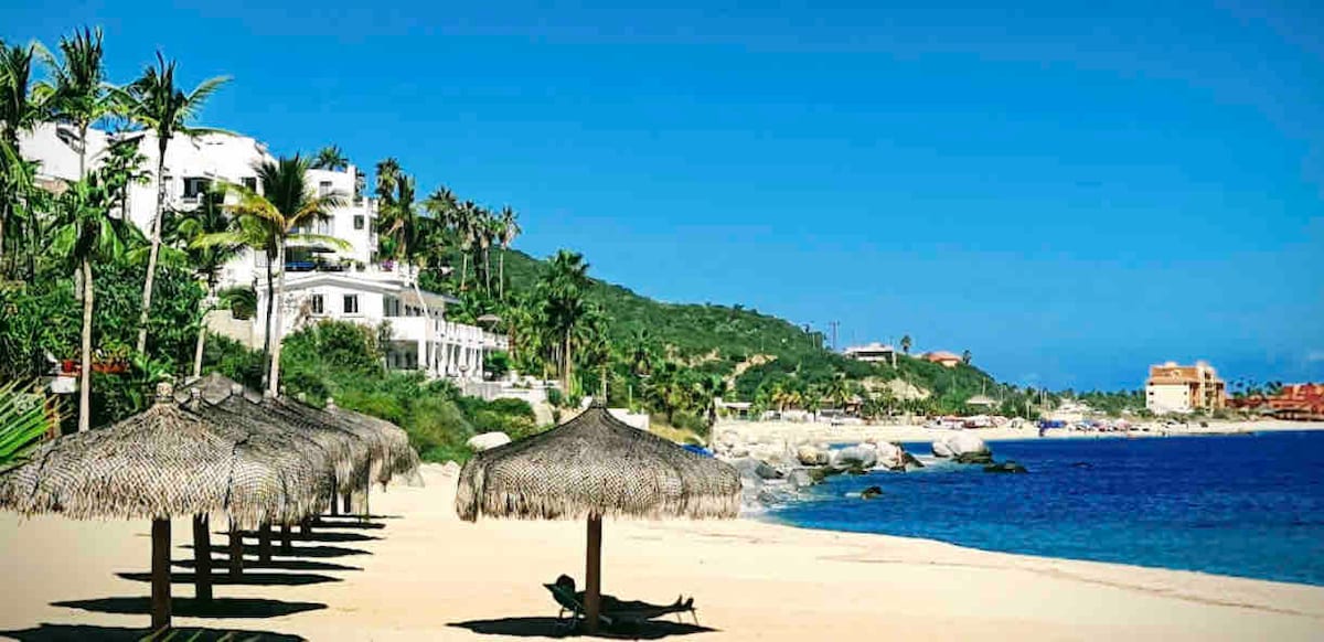 A sandy beach extends along the coastline, marked by several thatch-roof palapas providing shade. The bright blue ocean is visible, and lush palm trees line the beach, leading up to colorful buildings set against a green hillside in the distance.