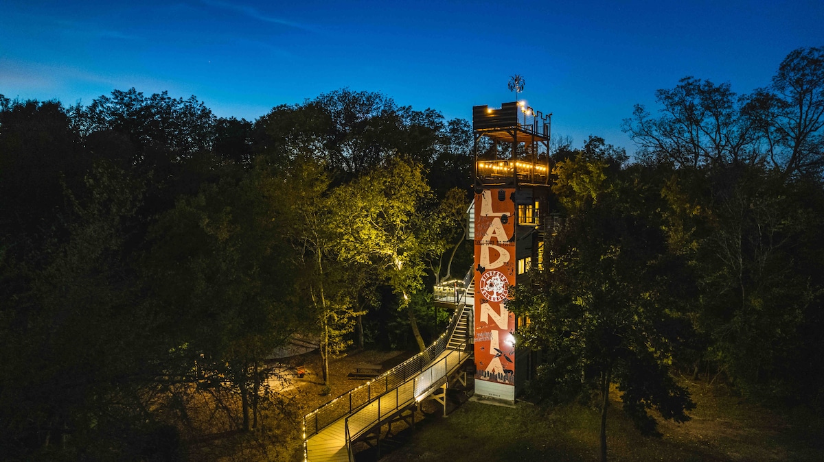Illuminated at night, the unique treehouse stands tall among surrounding trees. A wooden walkway leads to the entrance, adorned with vibrant artwork that adds character. Soft lighting highlights the structure against a deep blue sky, creating an inviting and serene atmosphere in nature.