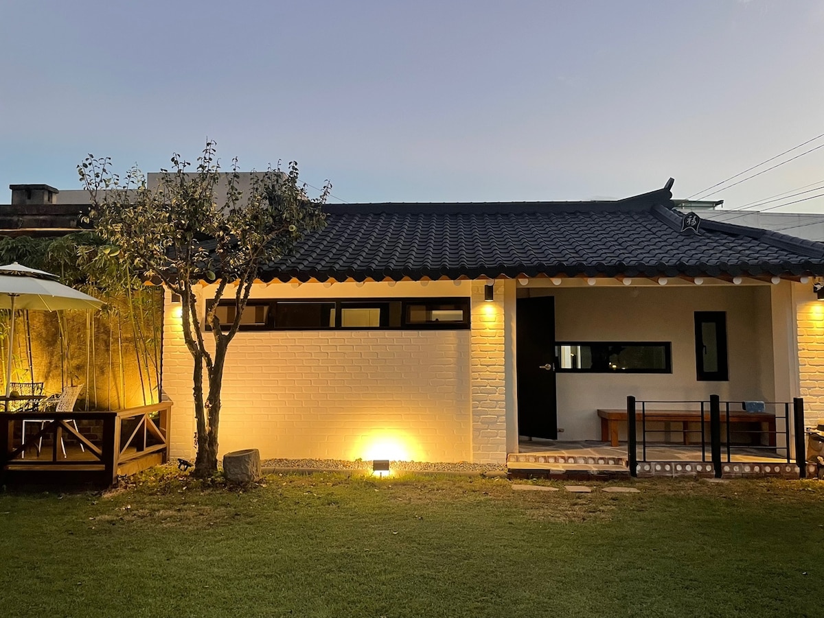 A traditional Korean house is set against a twilight sky. The exterior features white brick walls and a black tiled roof, illuminated softly by ground lights. A small bench is positioned near the entrance, with a well-maintained lawn and a sitting area visible in the background.