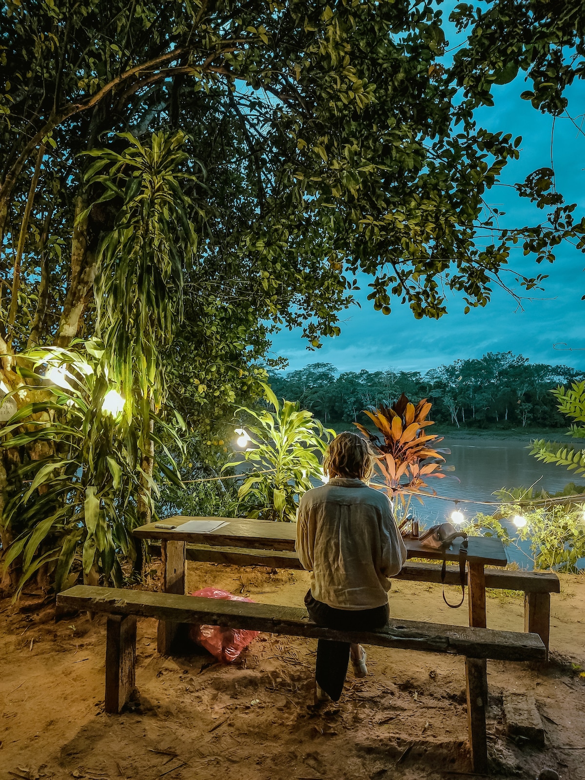A wooden table and benches are situated by the river, surrounded by lush greenery and softly illuminated by string lights. The tranquil river can be seen in the background, adding to the serene atmosphere of the setting.