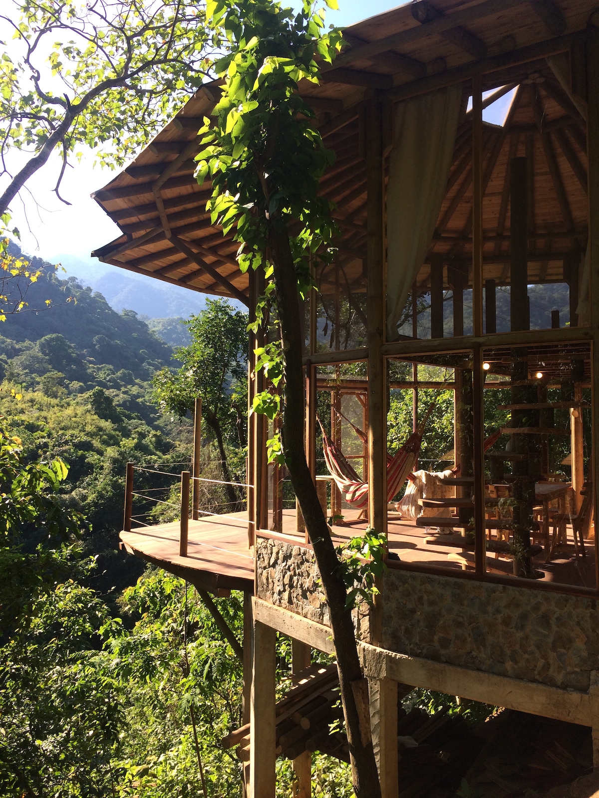 The wooden structure is elevated among lush greenery, featuring a spacious terrace adorned with a hammock. Large windows allow natural light to illuminate the living area. Surrounding mountains and dense jungle create a serene backdrop, enhancing the connection with nature.