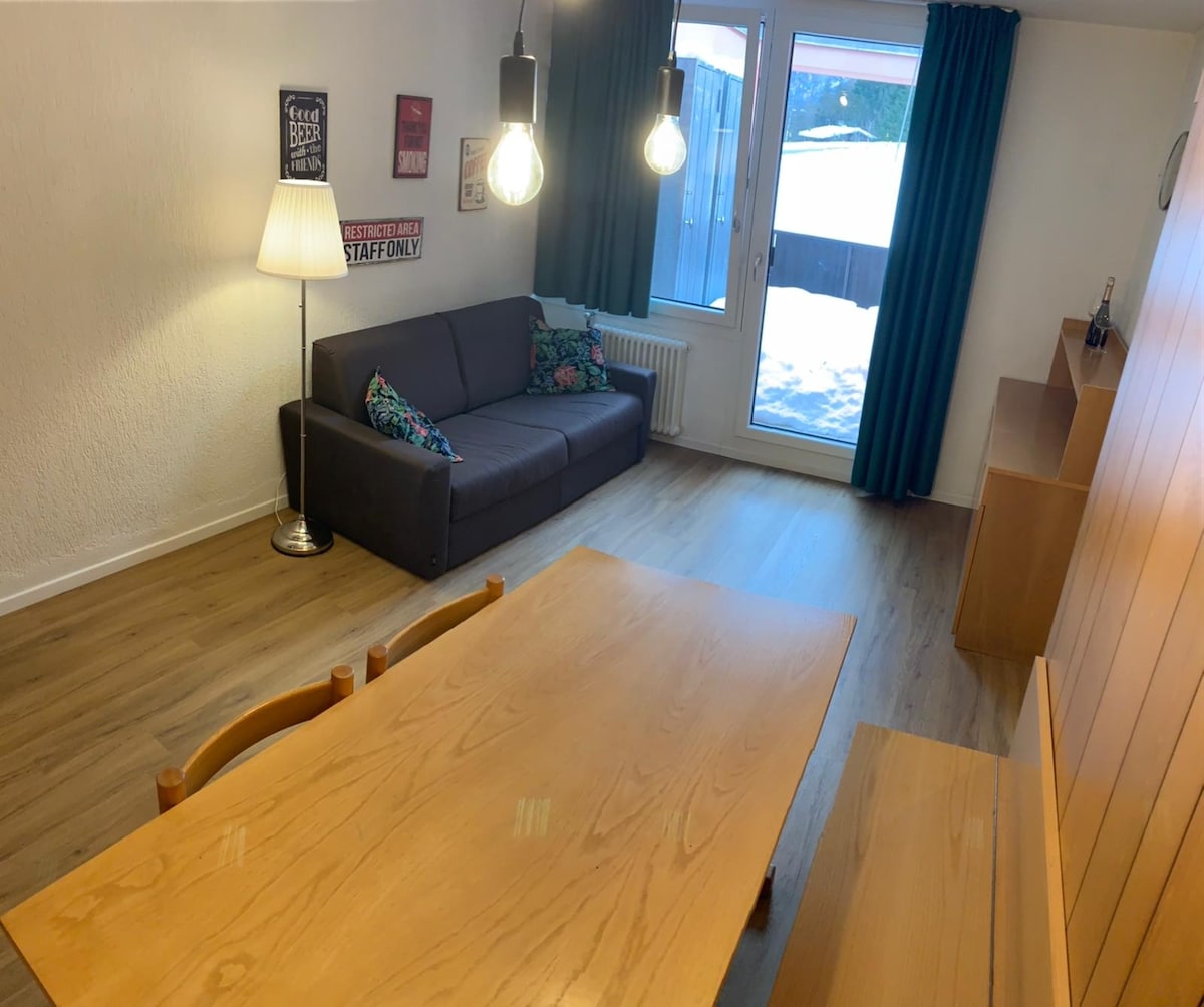 A spacious living area features a dark sofa adorned with colorful cushions and a wooden dining table. Large windows allow natural light to fill the room, with curtains that frame the view of the outdoors. A floor lamp adds a warm touch to the contemporary space.