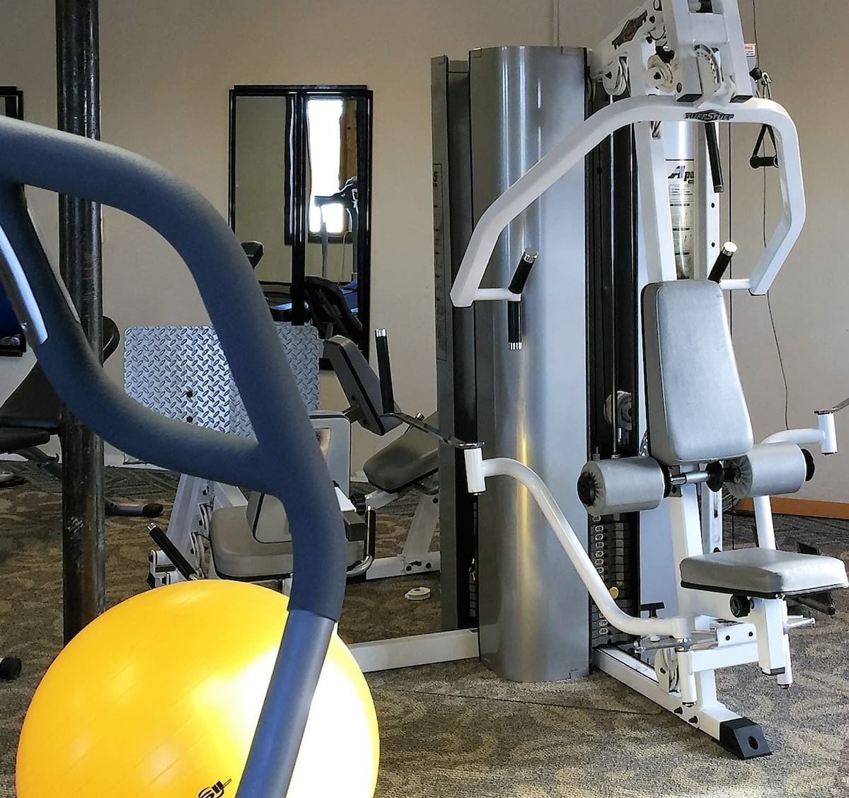 A fitness area is equipped with various exercise machines, including strength training equipment and a stability ball. The floor features a patterned carpet, and mirrors are positioned on the walls, enhancing the sense of space and allowing users to monitor their form.