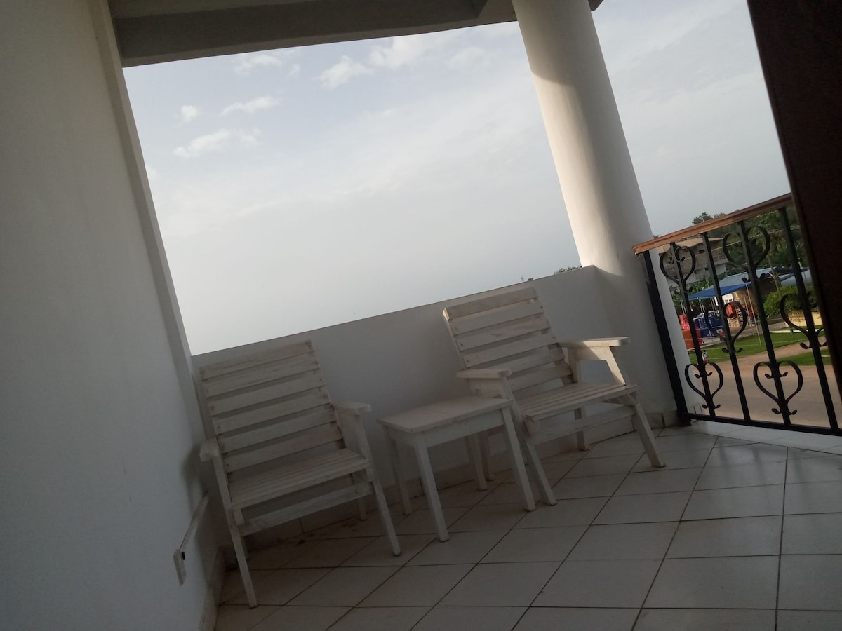 A small outdoor porch area is shown, featuring two wooden chairs and a side table. The space is surrounded by a white wall and offers a view of the outdoors beyond the railing, enhancing the sense of openness.