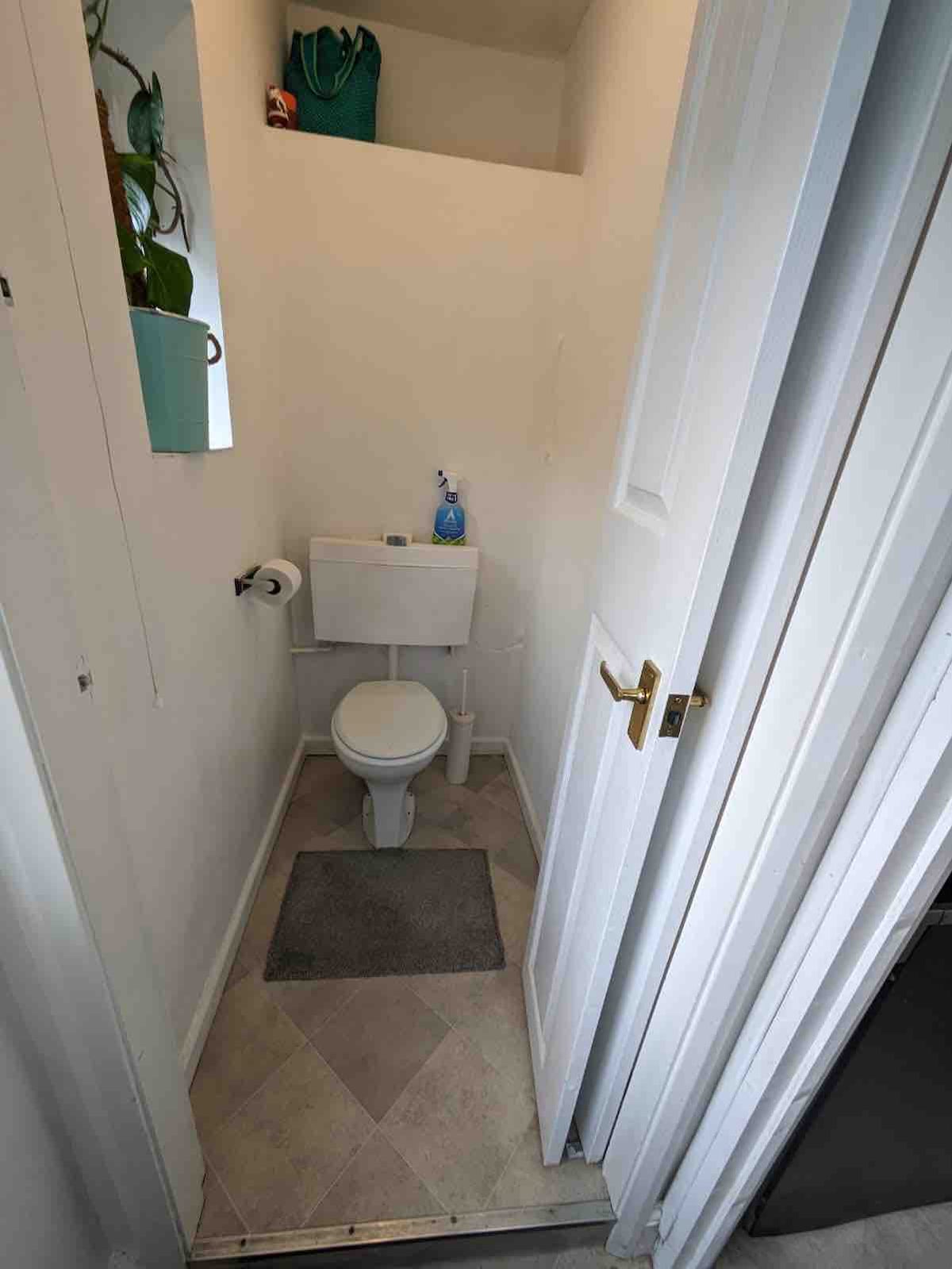 A small bathroom is displayed, featuring a toilet adjacent to a wall. A toilet paper holder is mounted next to it, and a floor mat is positioned on the tiled floor. Light enters from a small window above, enhancing the bright, clean space.