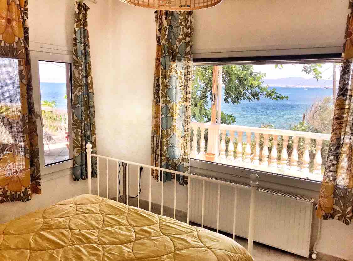 A bright bedroom features large windows offering views of the coastline. Colorful curtains frame the windows, while a quilted bedspread covers a spacious bed. Natural light fills the room, complemented by a simple light fixture hanging from the ceiling.