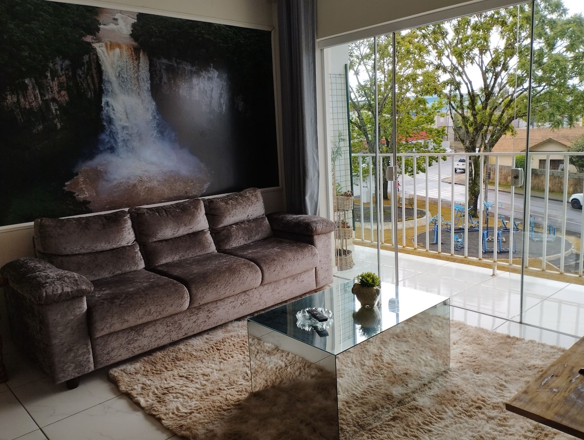 A comfortable living area features a plush gray sofa facing a large mural of a waterfall. A glass coffee table sits atop a light-colored rug, and natural light stream through the sliding glass doors that open to a balcony overlooking the street.
