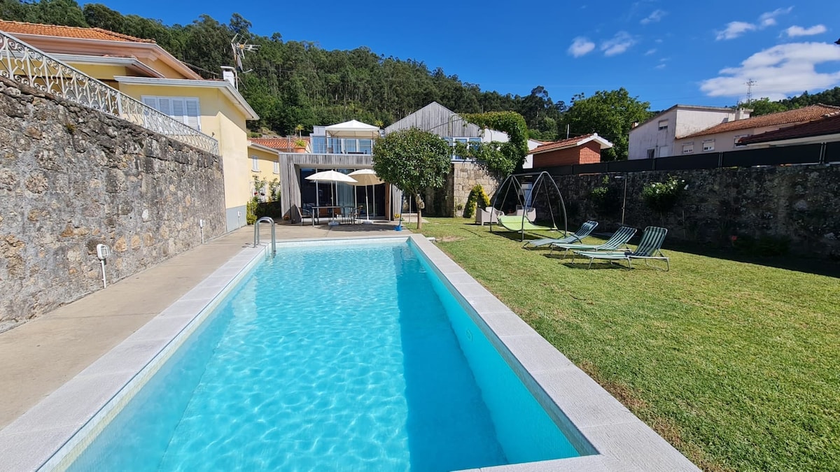 An outdoor area features a swimming pool with clear blue water, surrounded by a well-maintained lawn. Sun loungers are positioned nearby, and a covered seating area is visible in the background. A stone wall provides a backdrop, along with lush greenery from nearby trees.