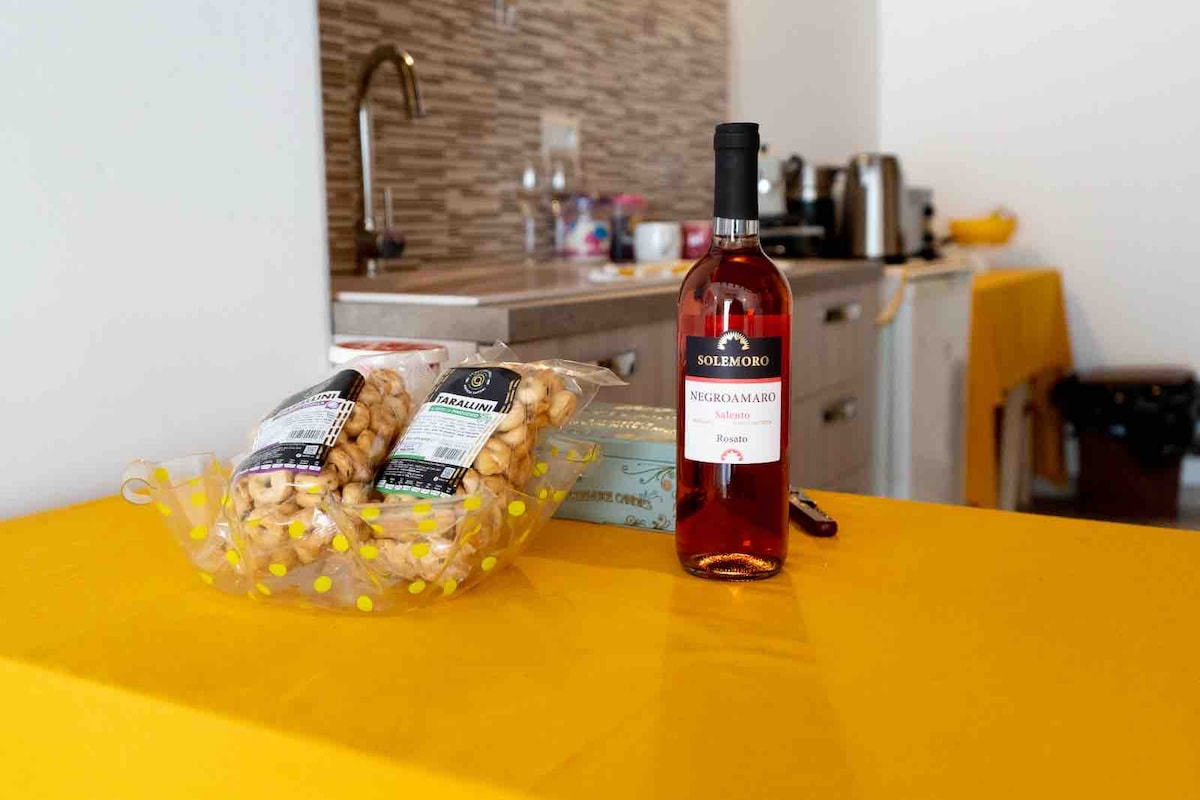A bright yellow table is set with a bottle of rosé wine and a selection of snacks, including packaged cookies and pastries. In the background, the compact kitchen features modern appliances and a subtle backsplash.