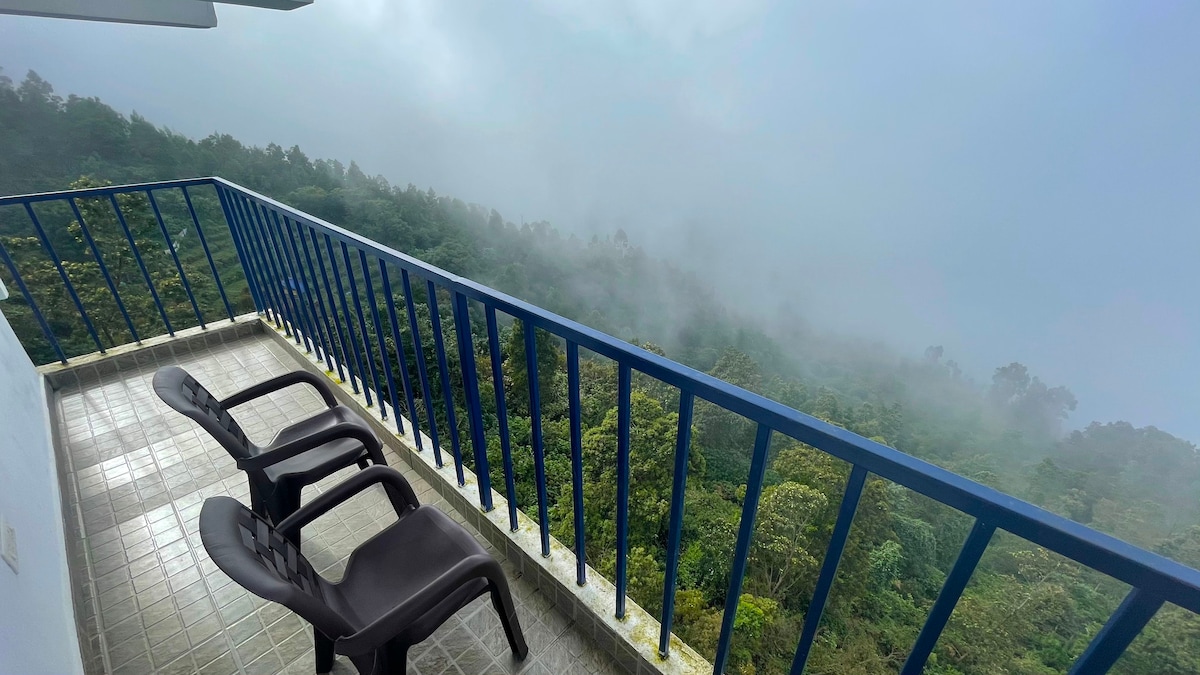 A balcony features two black lounge chairs, overlooking a lush green landscape enveloped in mist. The blue railings create a contrast against the serene backdrop, enhancing the feeling of openness and connection to nature.