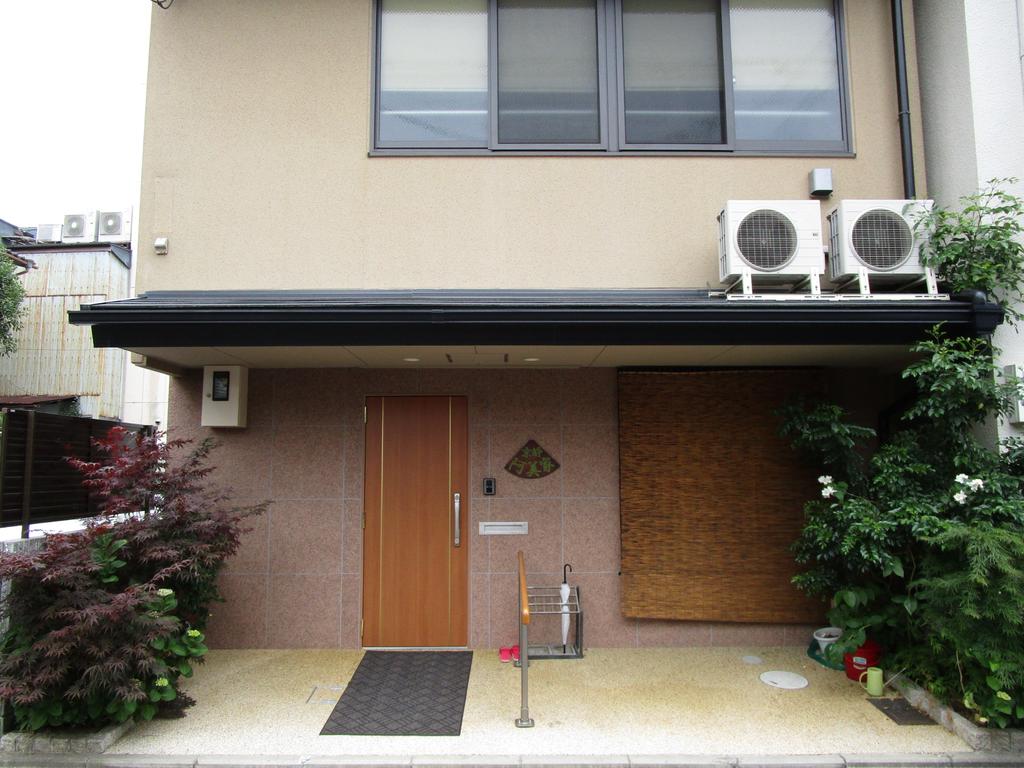 The entrance features a welcoming wooden door flanked by greenery and ornamental plants. A textured stone pathway leads to the door, while a covered area with air conditioning units is visible on the right. Natural elements complement the building's modern facade.