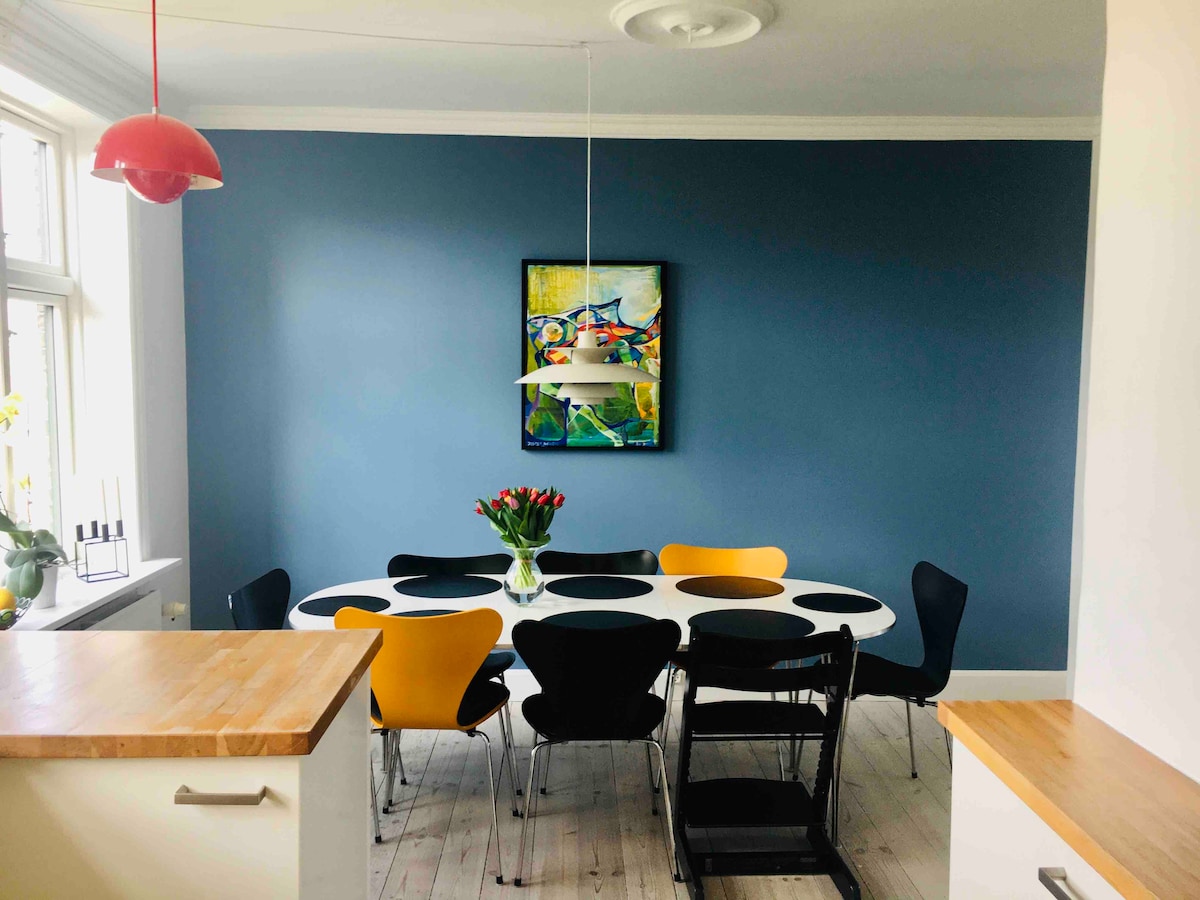 A spacious dining area is highlighted by a round table surrounded by colorful chairs in black and yellow. A vase of tulips sits at the center, and a vibrant piece of wall art adds a pop of color against the blue wall. Natural light enters through large windows.