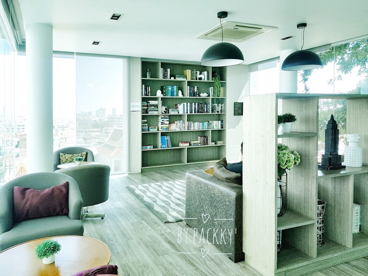 A modern living area is displayed, featuring a cozy sofa and two curved chairs. A well-organized bookshelf filled with various items is visible in the background. Large windows allow natural light to illuminate the space, providing a view of the city outside.