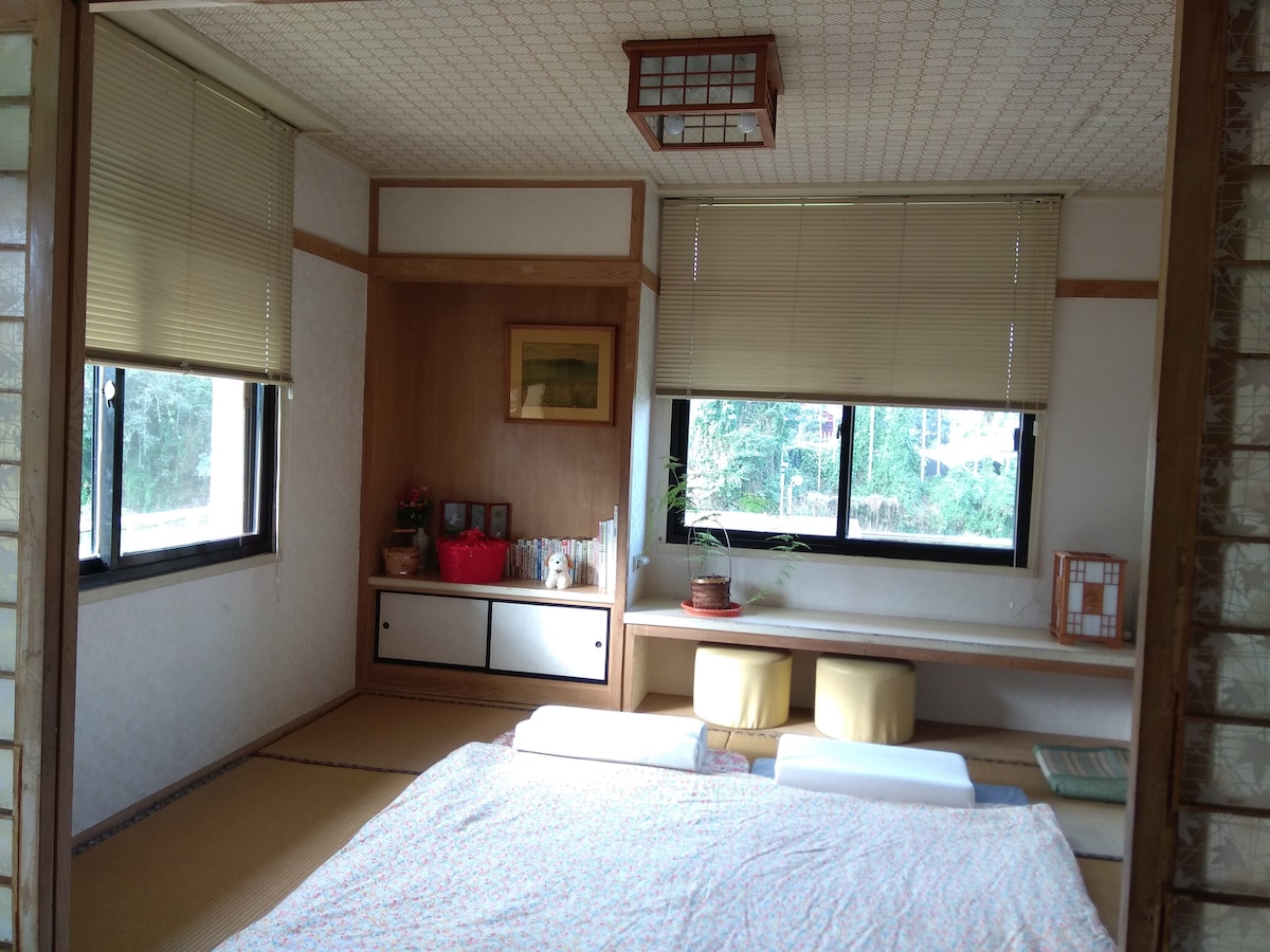 A serene tatami room is featured, with natural light filtering through two large windows. The space includes a low seating area with two stools and wall shelves displaying decor. A spacious sleeping area is visible with bedding laid out on the tatami floor.