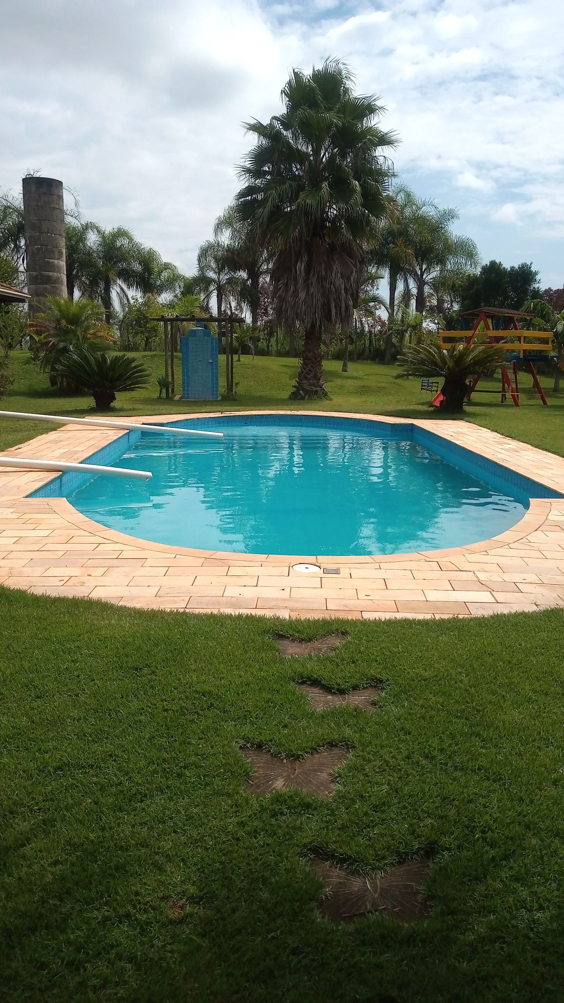 A sparkling blue swimming pool is framed by a tiled patio, surrounded by lush green grass. Palm trees and playground equipment can be seen in the background, creating a serene outdoor space suitable for families and events.