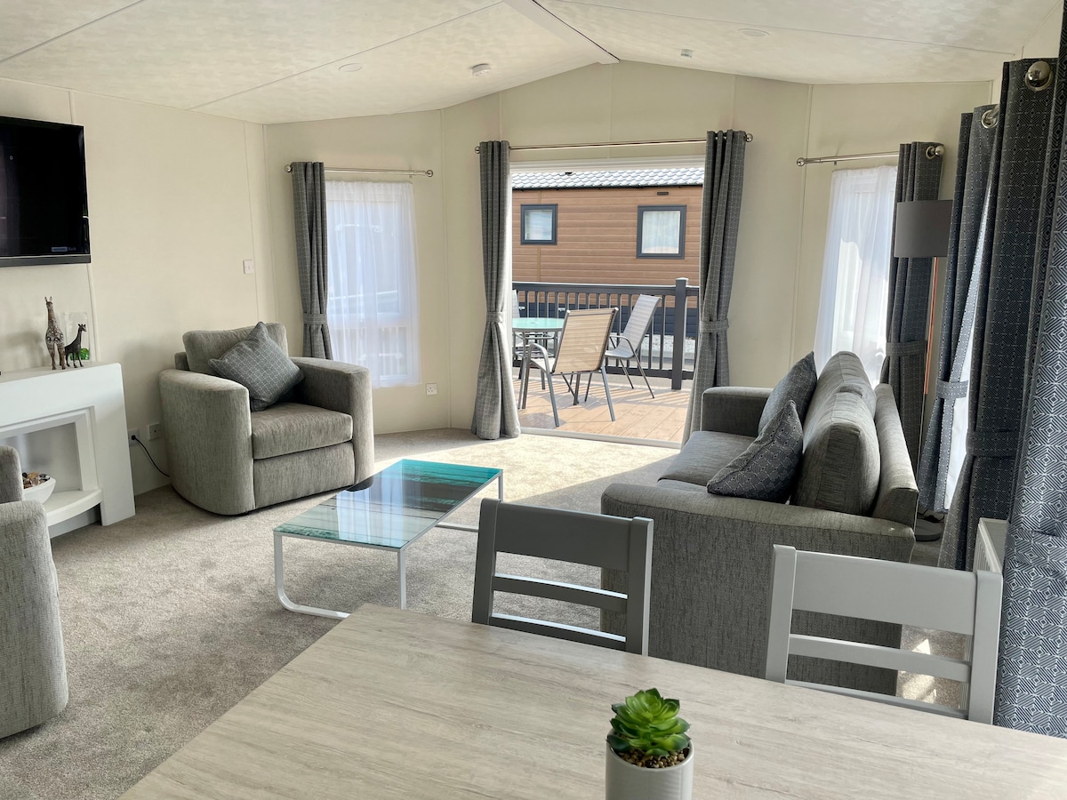 An open plan living area is depicted, featuring a comfortable seating arrangement with a sofa and armchair. A modern glass coffee table is centered on a neutral carpet, and large windows allow natural light to fill the space. A dining table with chairs is visible in the foreground.