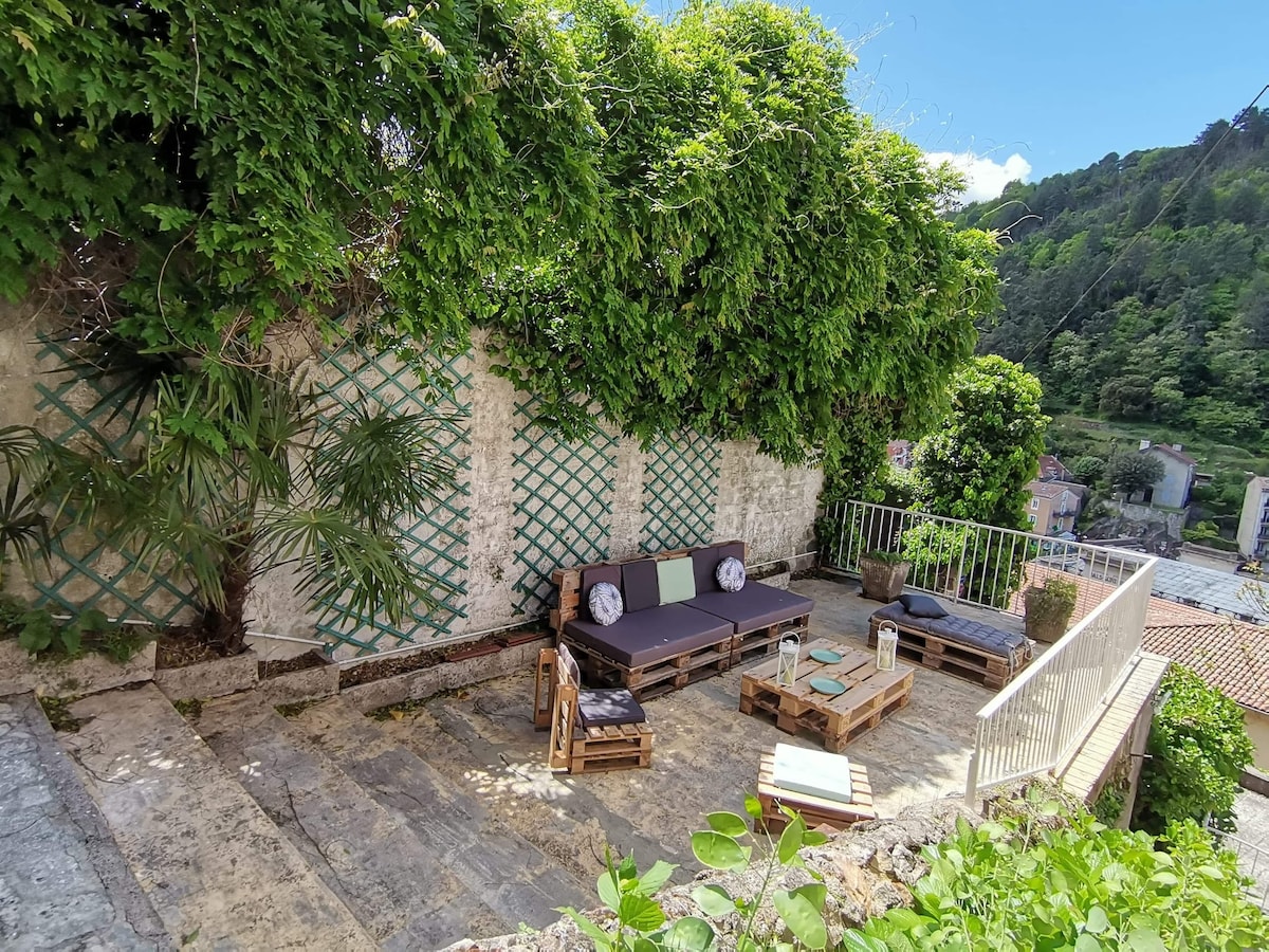 An outdoor terrace is surrounded by lush greenery and features multiple seating areas with comfortable cushions. Pallet furniture is arranged to create inviting spaces for relaxation, complemented by a small table. The scenic backdrop includes views of the hillside and nearby structures.