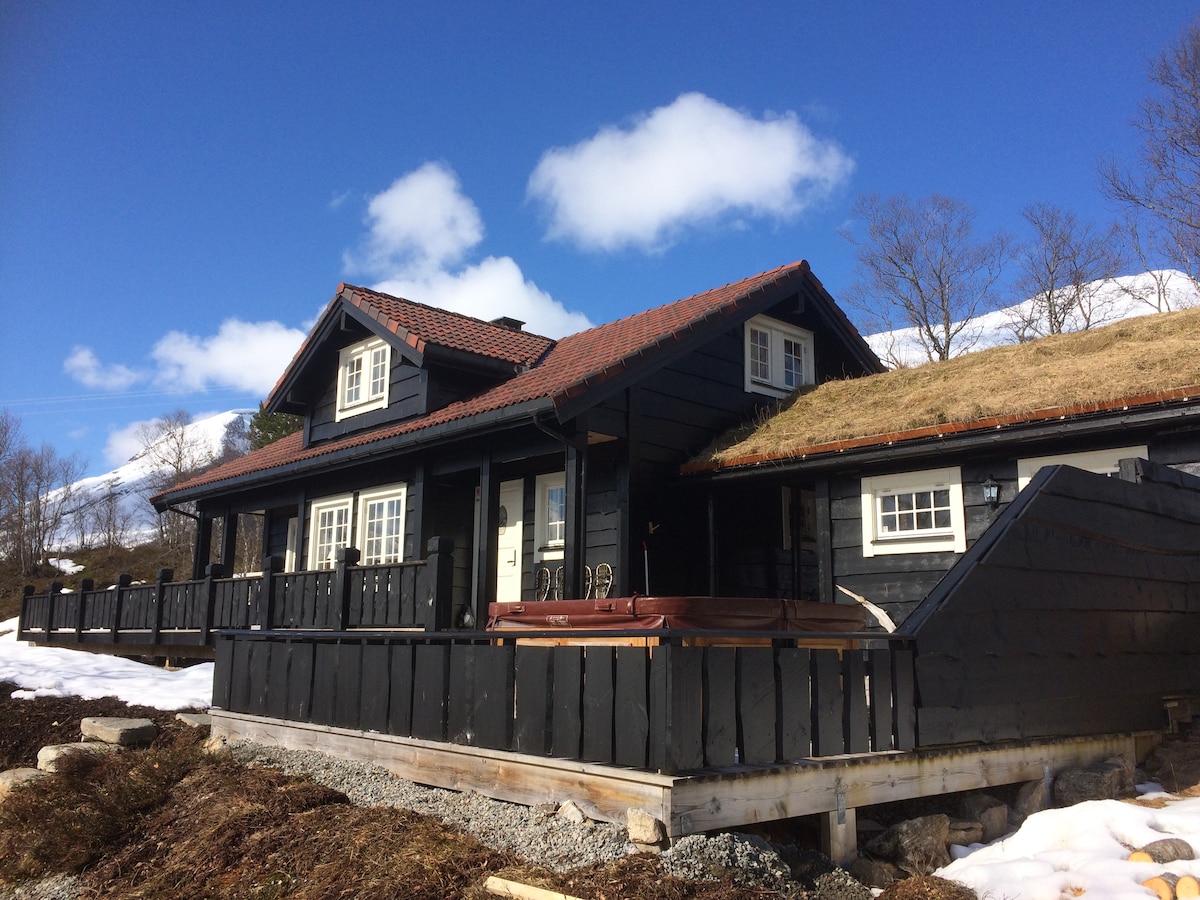 A chalet-style cabin features dark wood siding and a distinctive grass-covered roof. A spacious deck wraps around the cabin, providing outdoor seating. Snow covers the ground, contrasting with the clear blue sky, while trees are visible in the background.