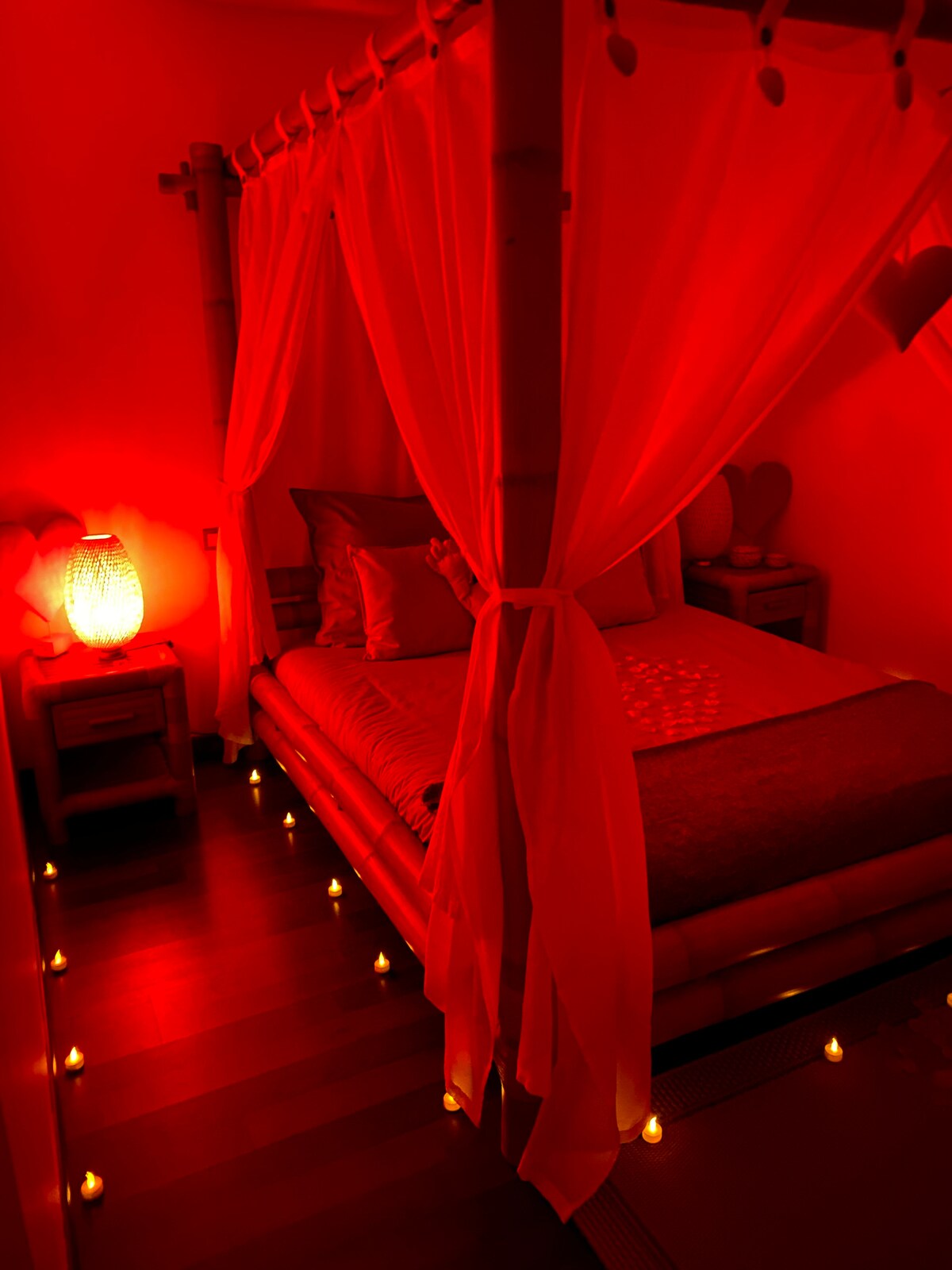 A cozy bedroom is softly illuminated with red lighting, creating a romantic ambiance. A spacious bed draped with sheer curtains is positioned centrally, surrounded by small candles that add to the serene atmosphere. Nightstands with lamps complement the decor.