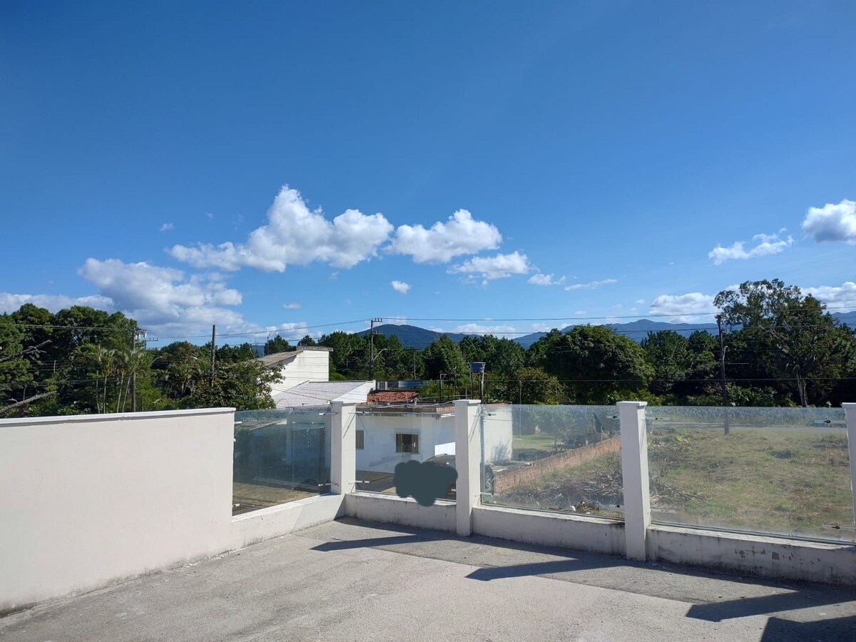 A rooftop terrace is shown, featuring a low glass barrier that allows for an unobstructed view of the surrounding greenery and distant mountains under a clear blue sky. The space offers ample sunlight, creating an open and airy atmosphere.