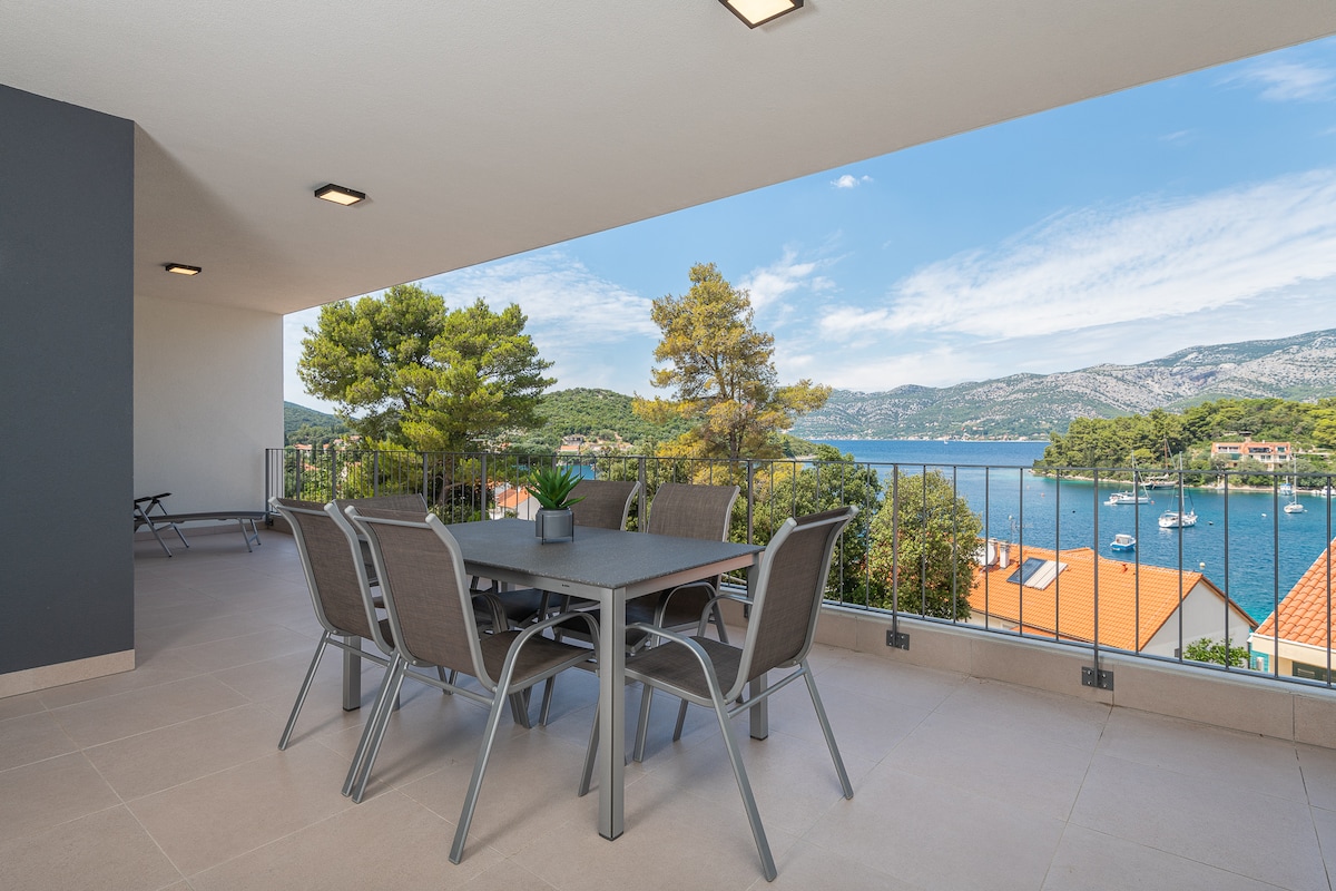 An outdoor balcony area features a spacious dining table surrounded by six grey chairs. Lush greenery and coastal views of the sea and surrounding hills create a serene backdrop. The sky is blue with scattered clouds, enhancing the relaxing atmosphere.