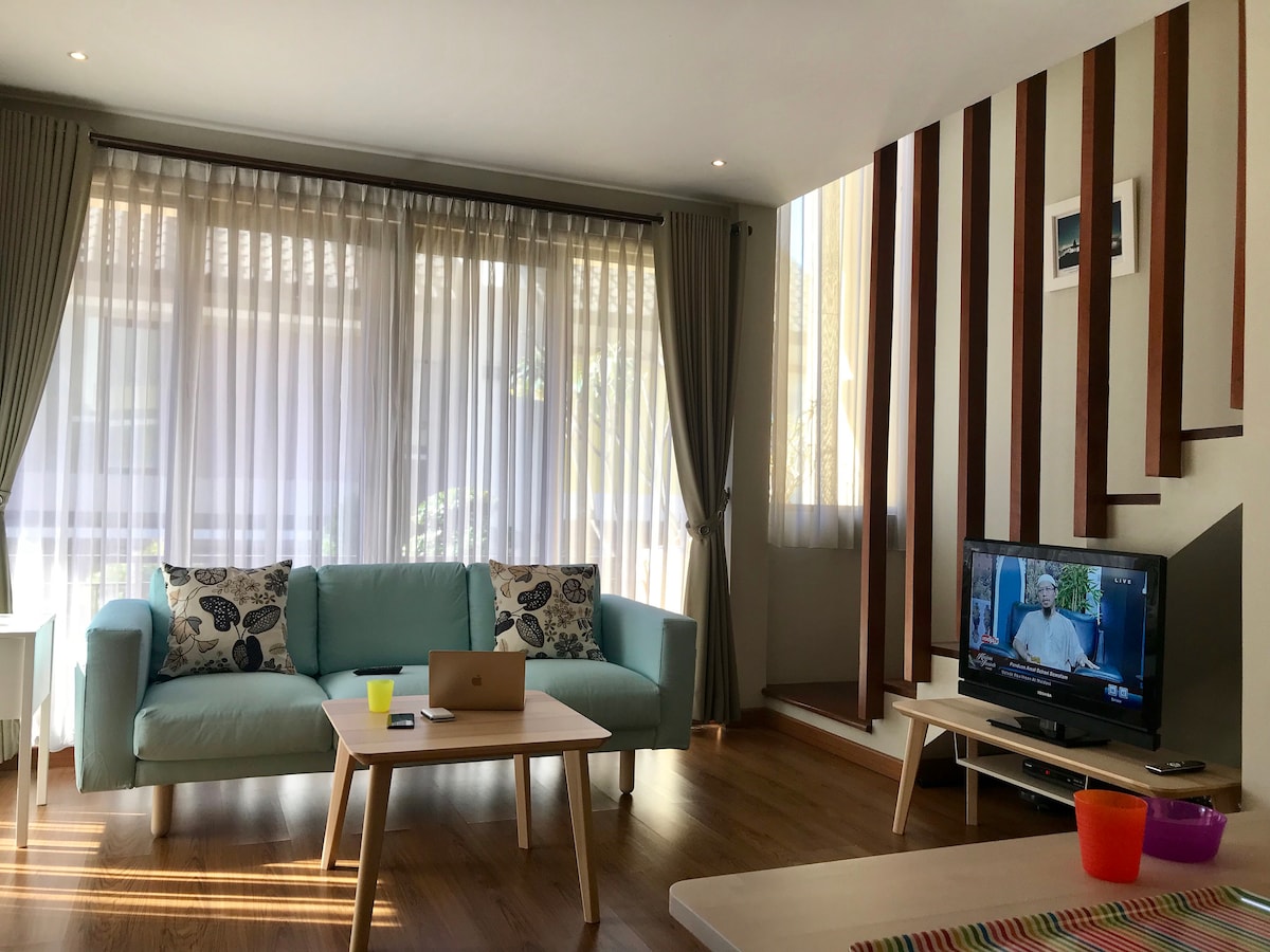 A well-lit living area features a light blue sofa adorned with decorative pillows. There are large windows with sheer curtains allowing natural light. A wooden coffee table is positioned in front of the sofa, while a television is placed opposite, showcasing a calm ambiance.