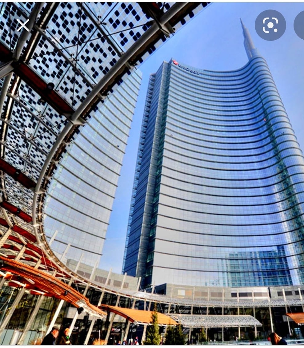 A modern skyscraper with a sleek glass exterior rises prominently, reflecting the sky above. The structure features a distinctive curved design at its base, framed by a canopy that showcases a blend of natural materials and contemporary architecture.