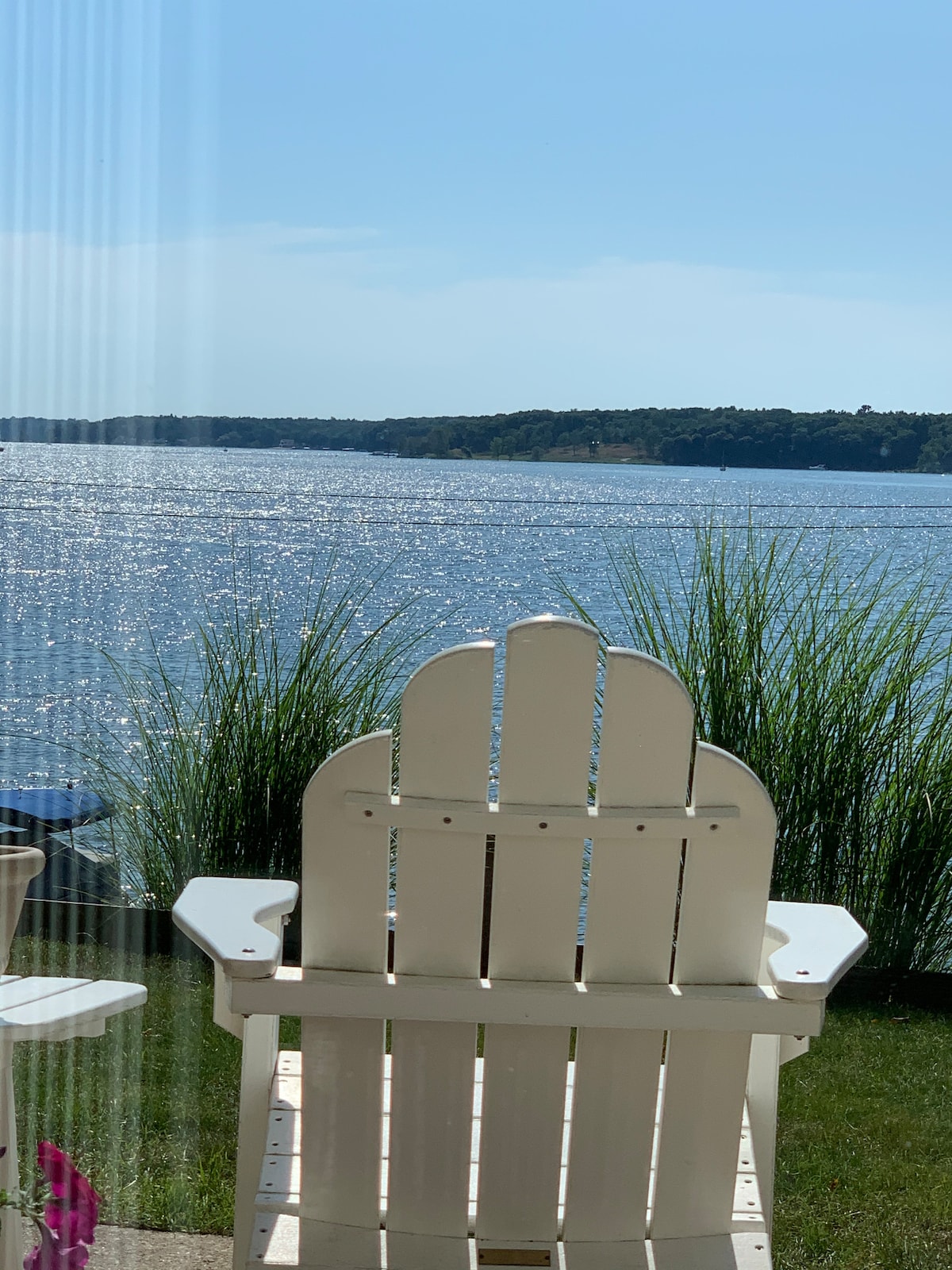 A white Adirondack chair is positioned on a patio, facing a shimmering expanse of water. Tall grasses frame the view, which includes distant tree-lined shores under a clear blue sky.